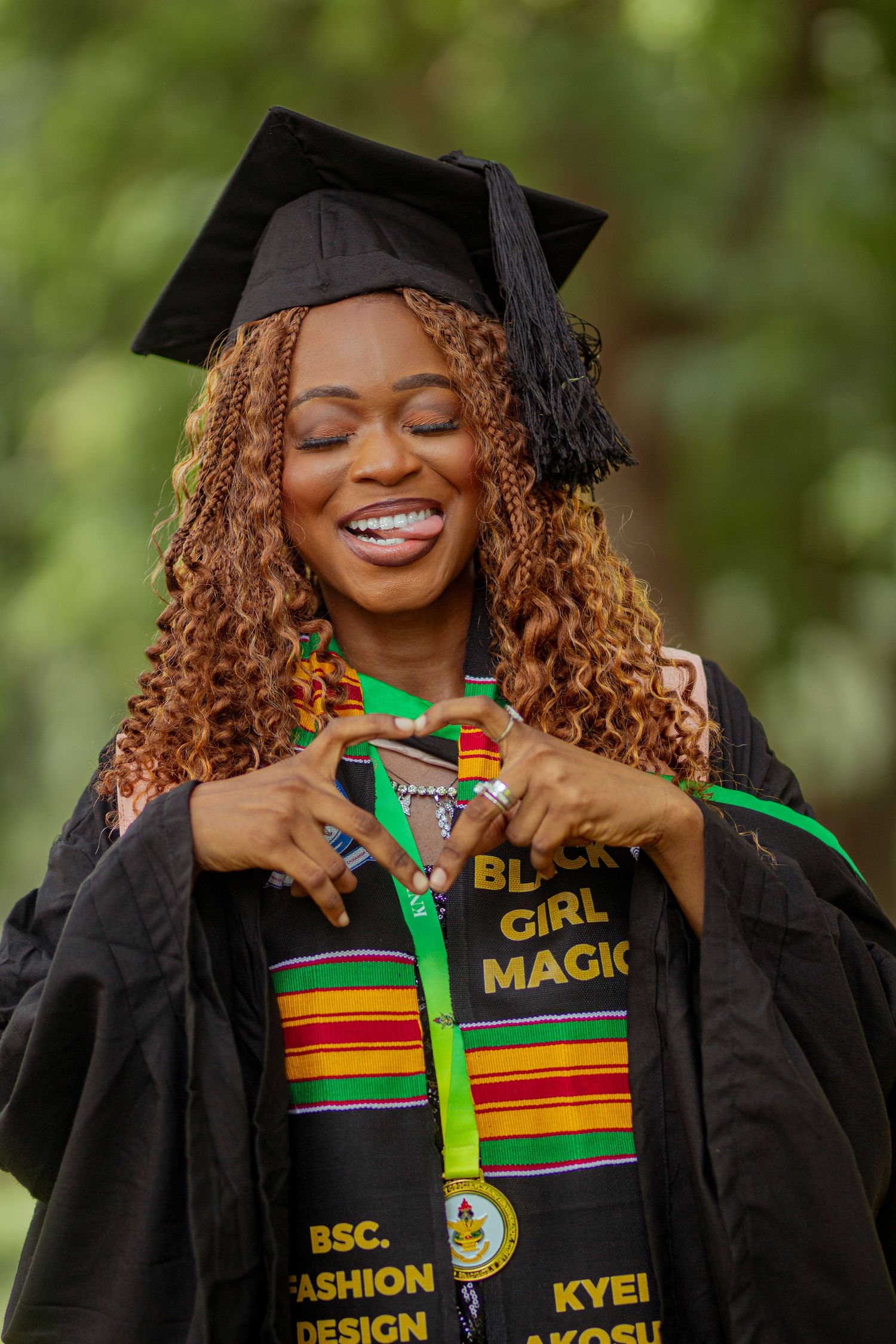 Graduate in black cap and gown with colorful kente stole makes heart shape gesture while smiling during graduation ceremony.