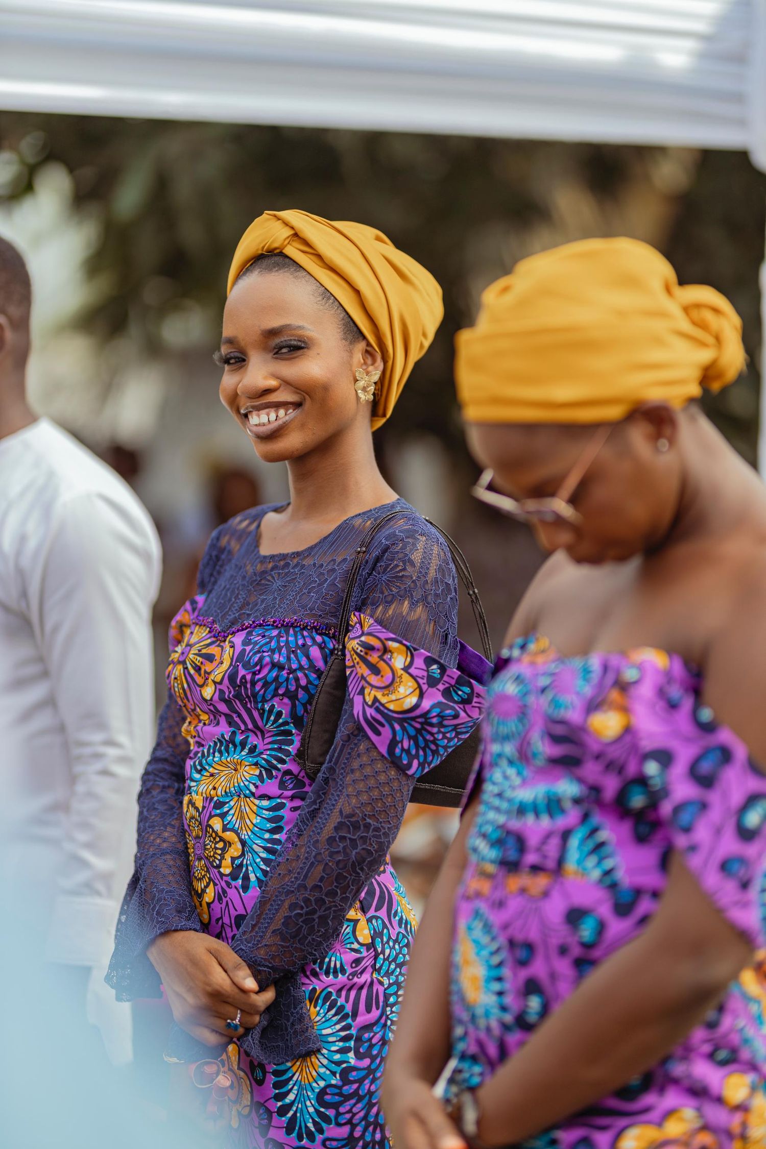 Guests in vibrant African print dresses and yellow head wraps attend a celebration outdoors.