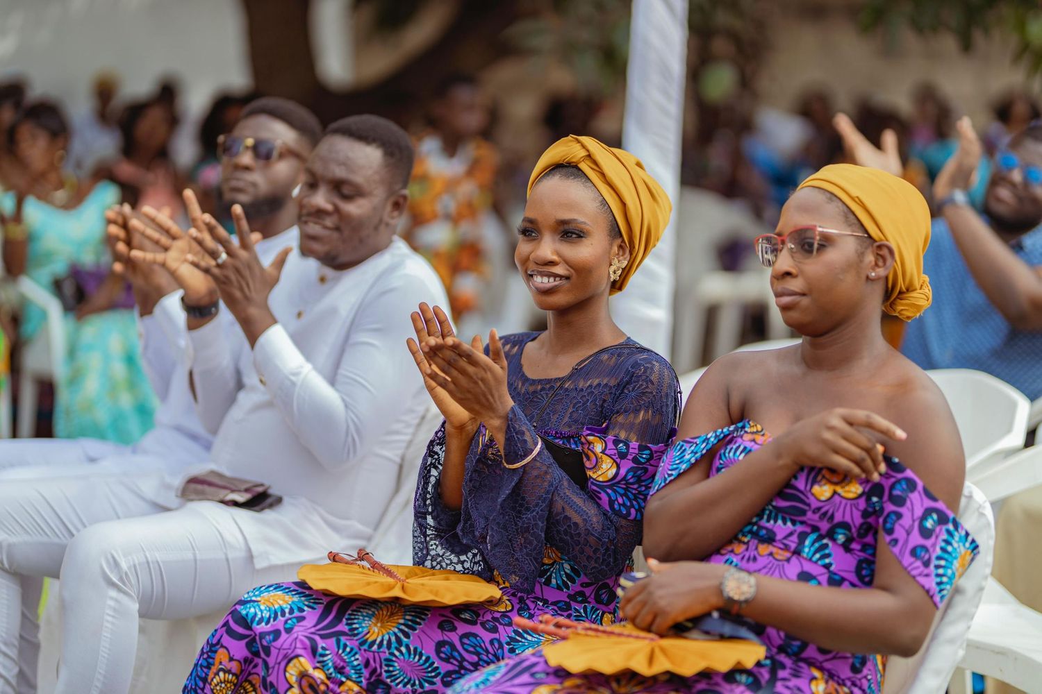 Wedding guests in colorful traditional African attire applaud during an outdoor celebration.