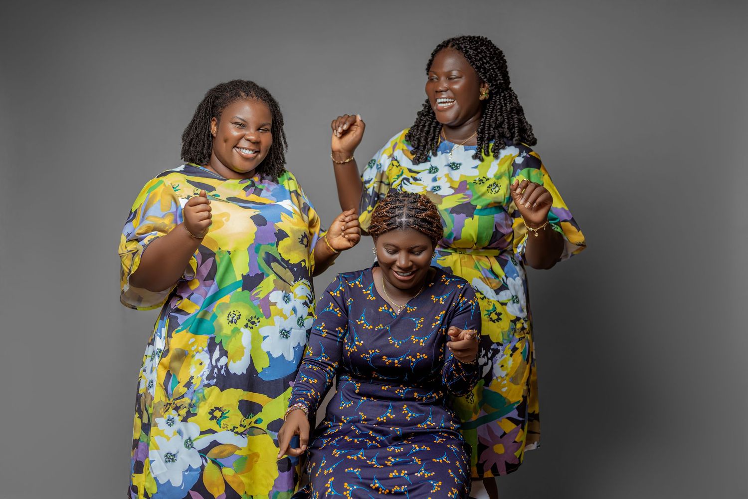 Group wearing vibrant floral print dresses poses joyfully against gray background.