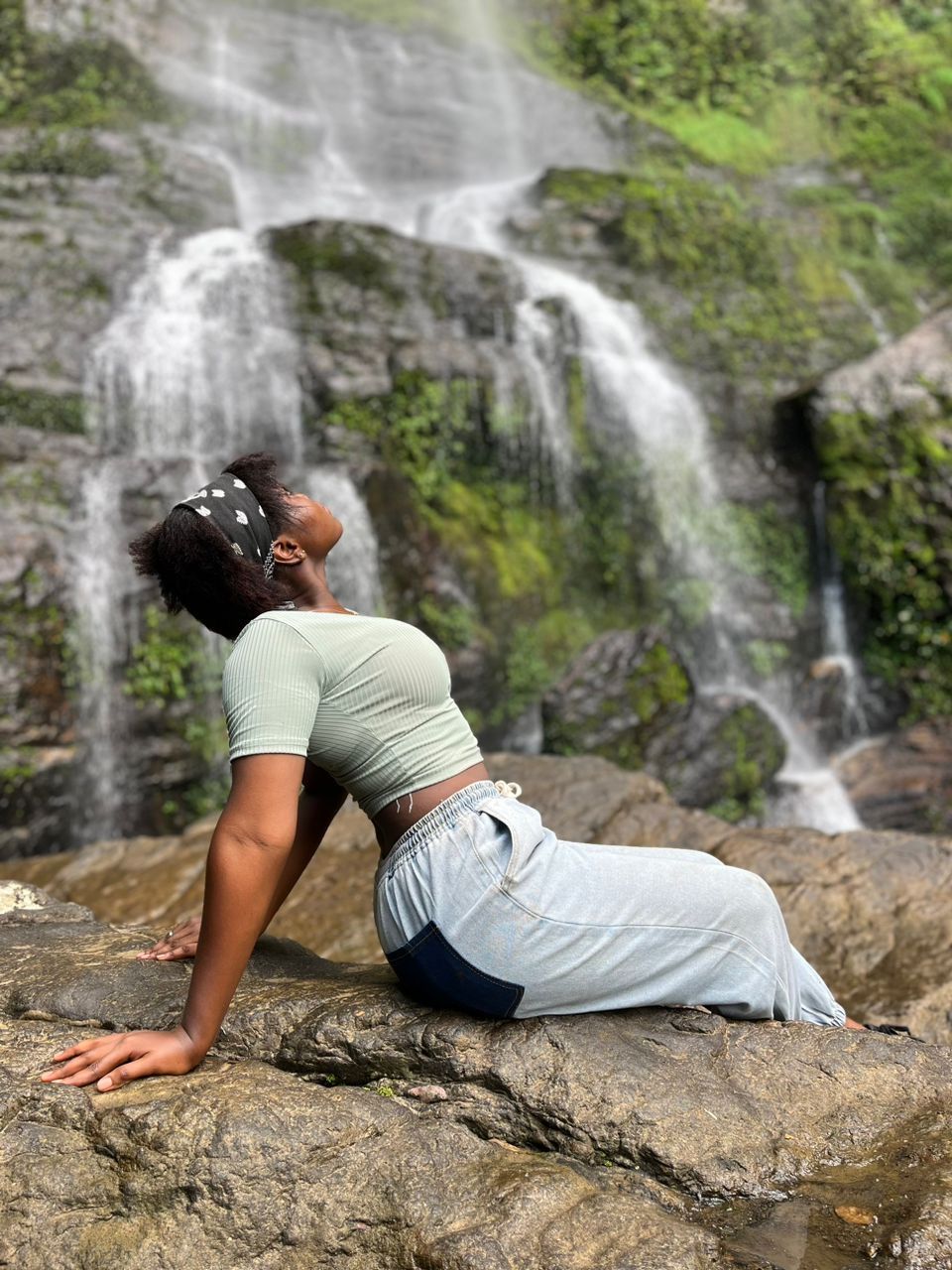 A person in casual attire relaxes on a rock in front of a scenic waterfall.