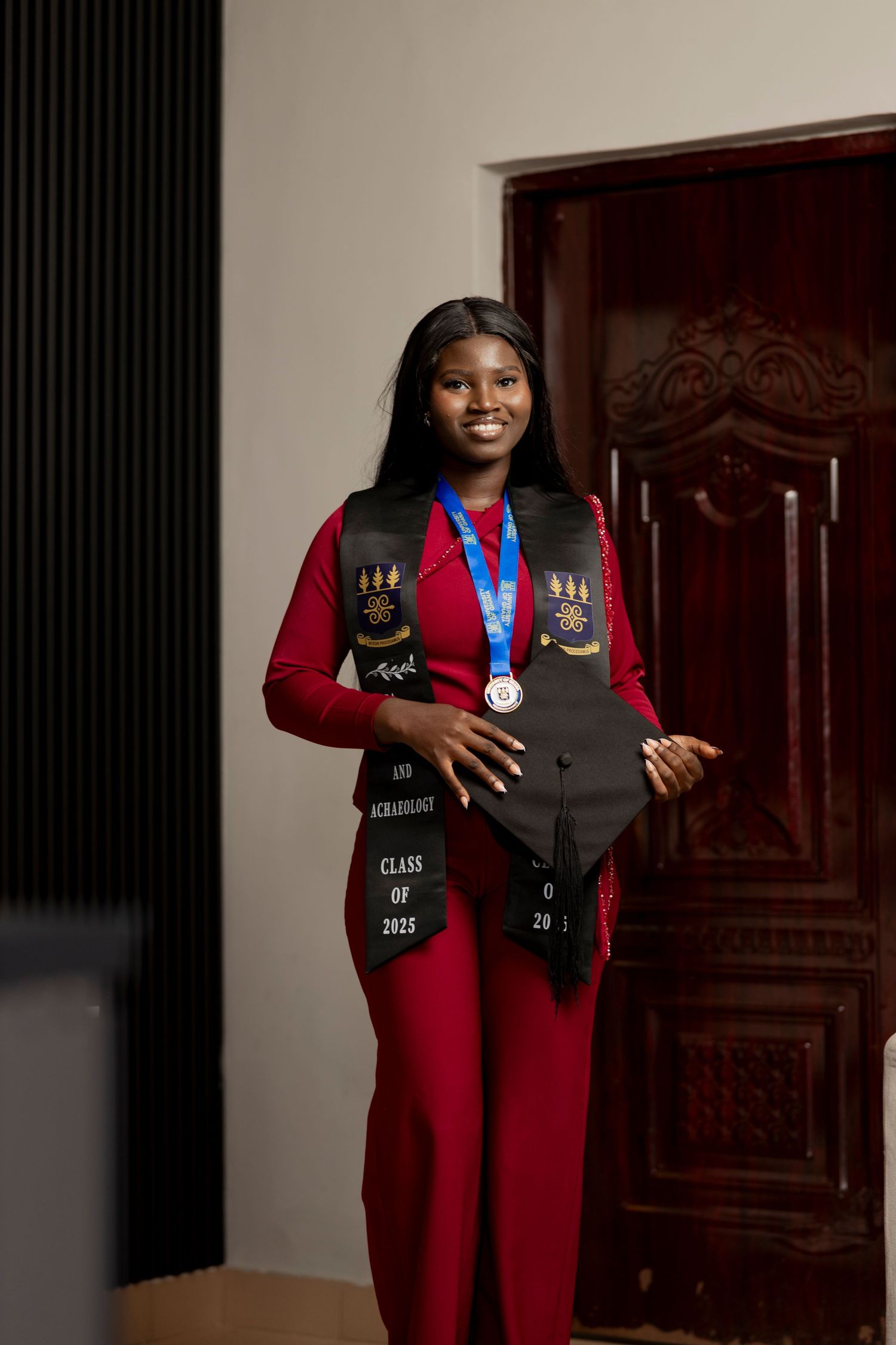 Smiling graduate in red outfit wearing black graduation stole, medal, and holding cap, Class of 2022.