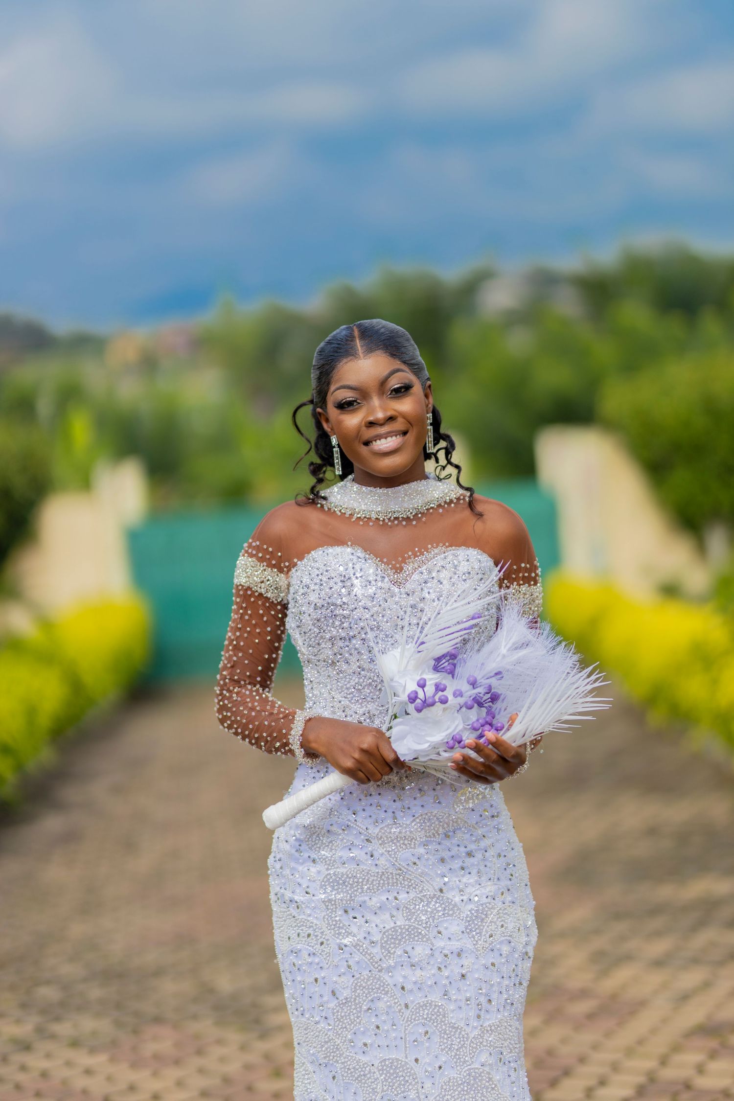 A bride in a sparkly white dress with mesh sleeves poses outdoors against a garden backdrop holding a white feather fan.