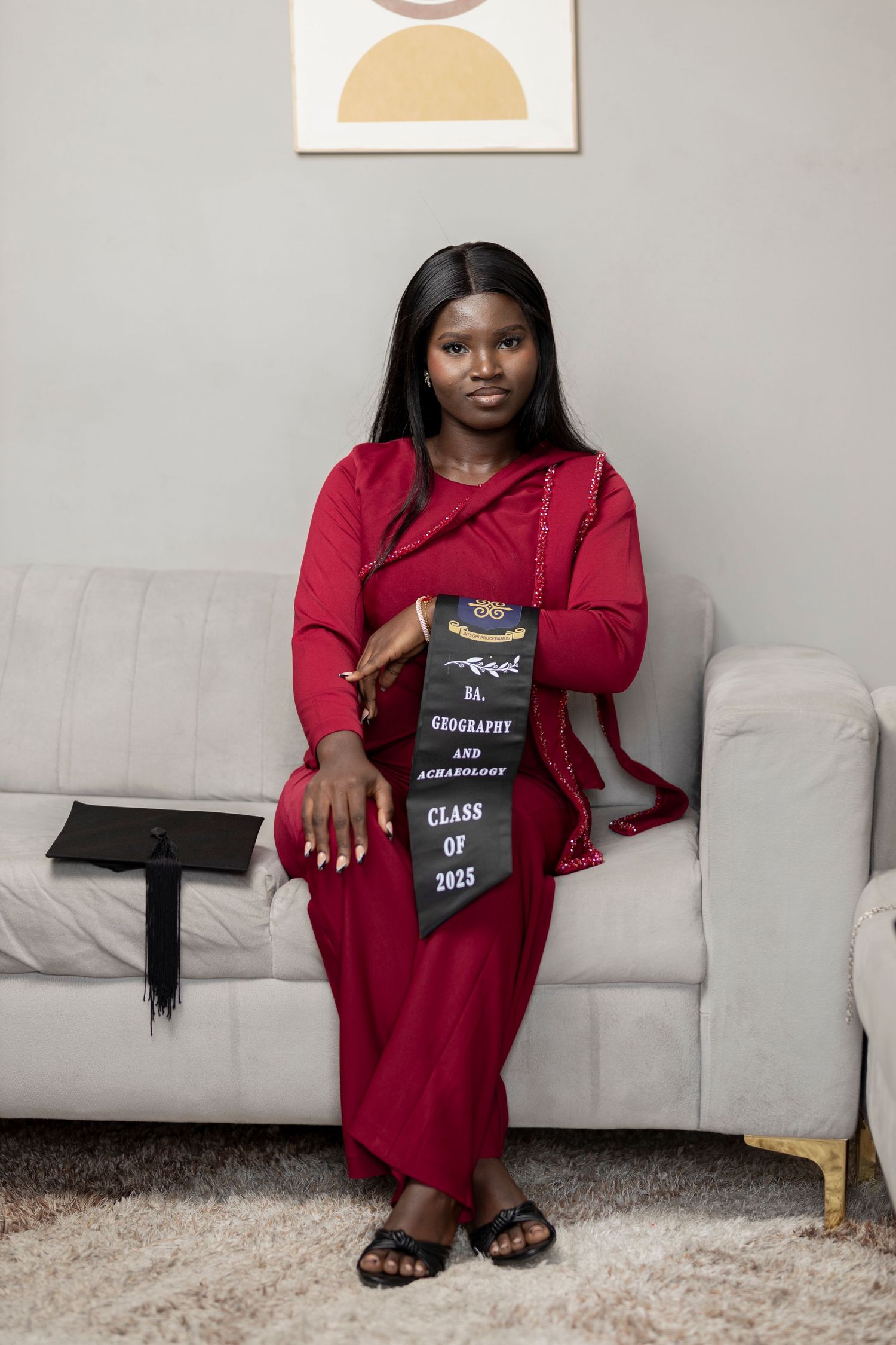 Young woman in red dress holding Class of 2025 Geography and Archaeology graduation stole, seated on white sofa.