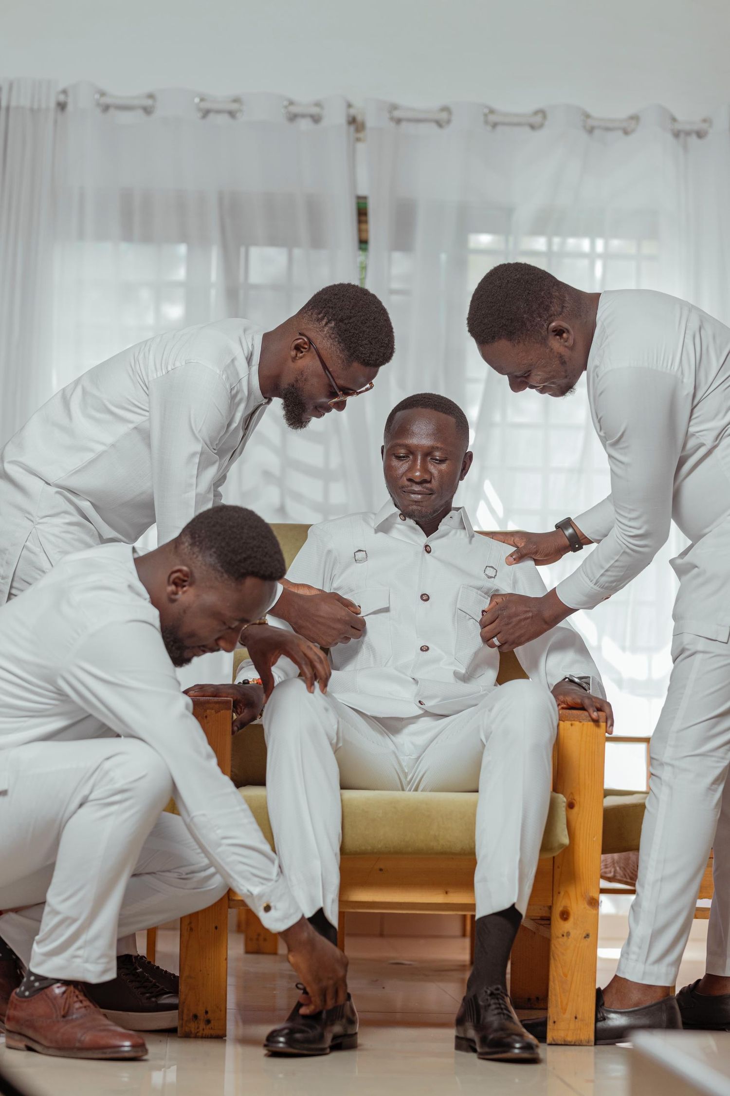 Four people in white formal wear gather closely around seated person during event.