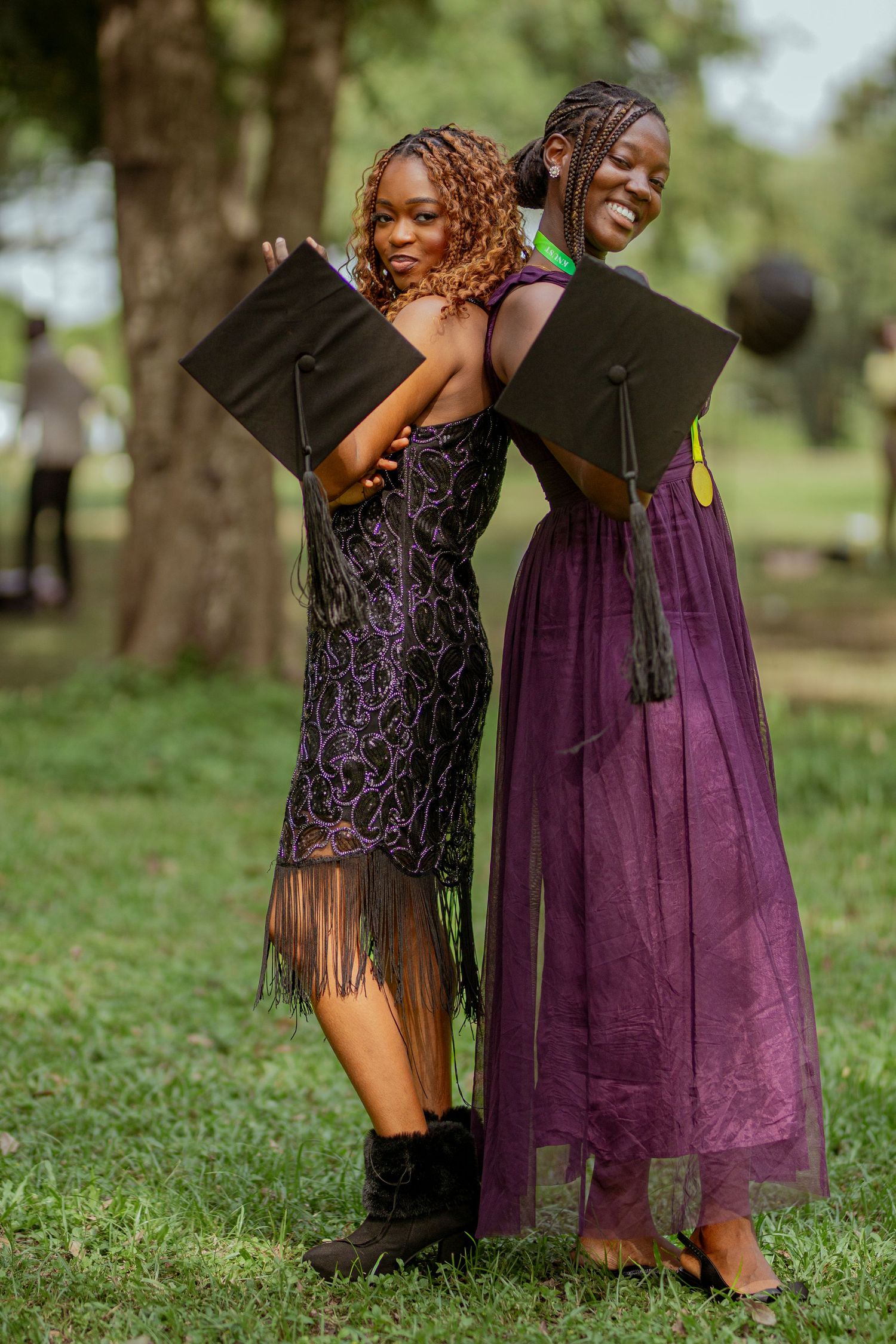 Two graduates in elegant dresses pose back-to-back while holding their mortarboards in an outdoor garden setting.