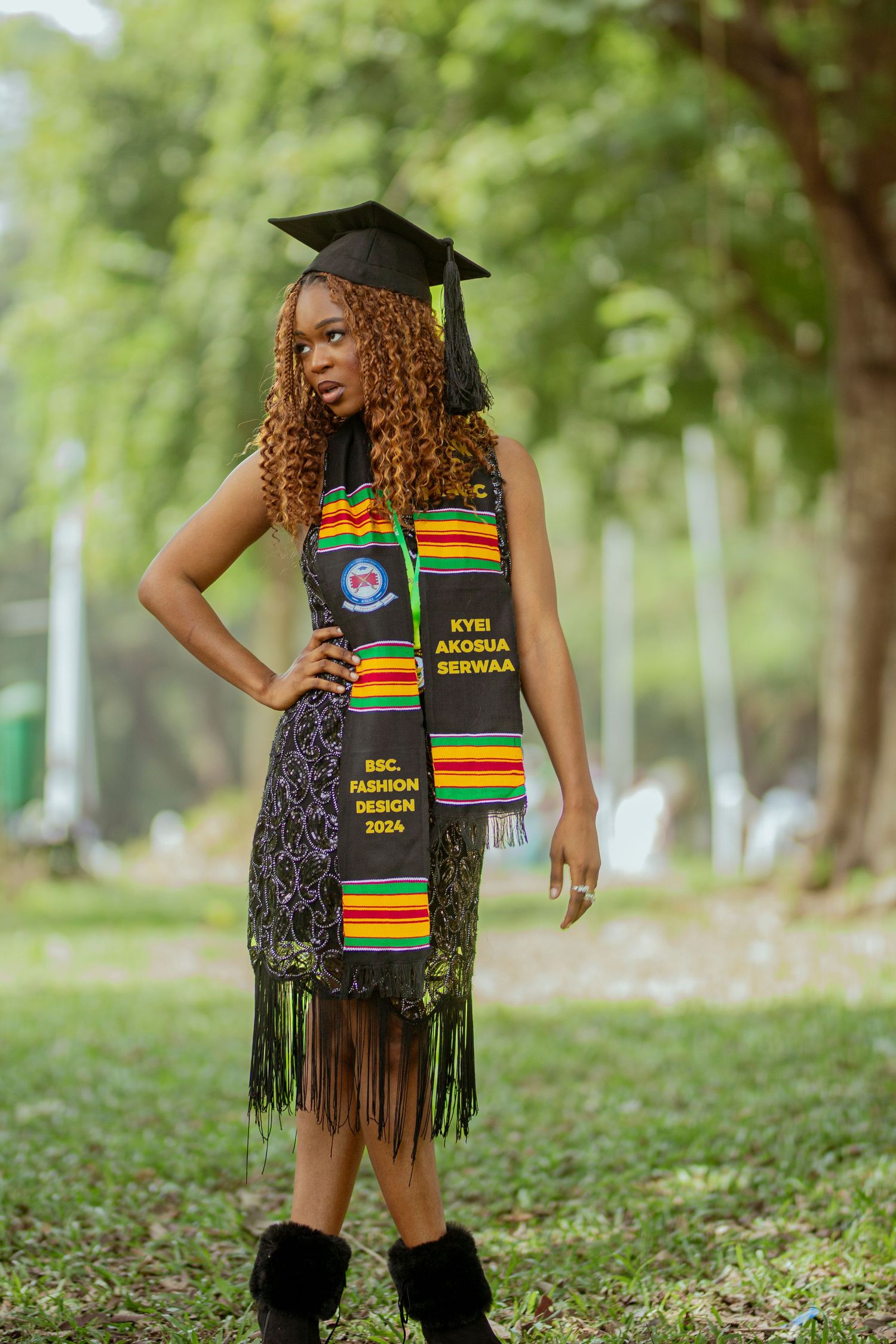 Graduate wearing traditional kente stole and black cap poses outdoors in grassy area for graduation photos.