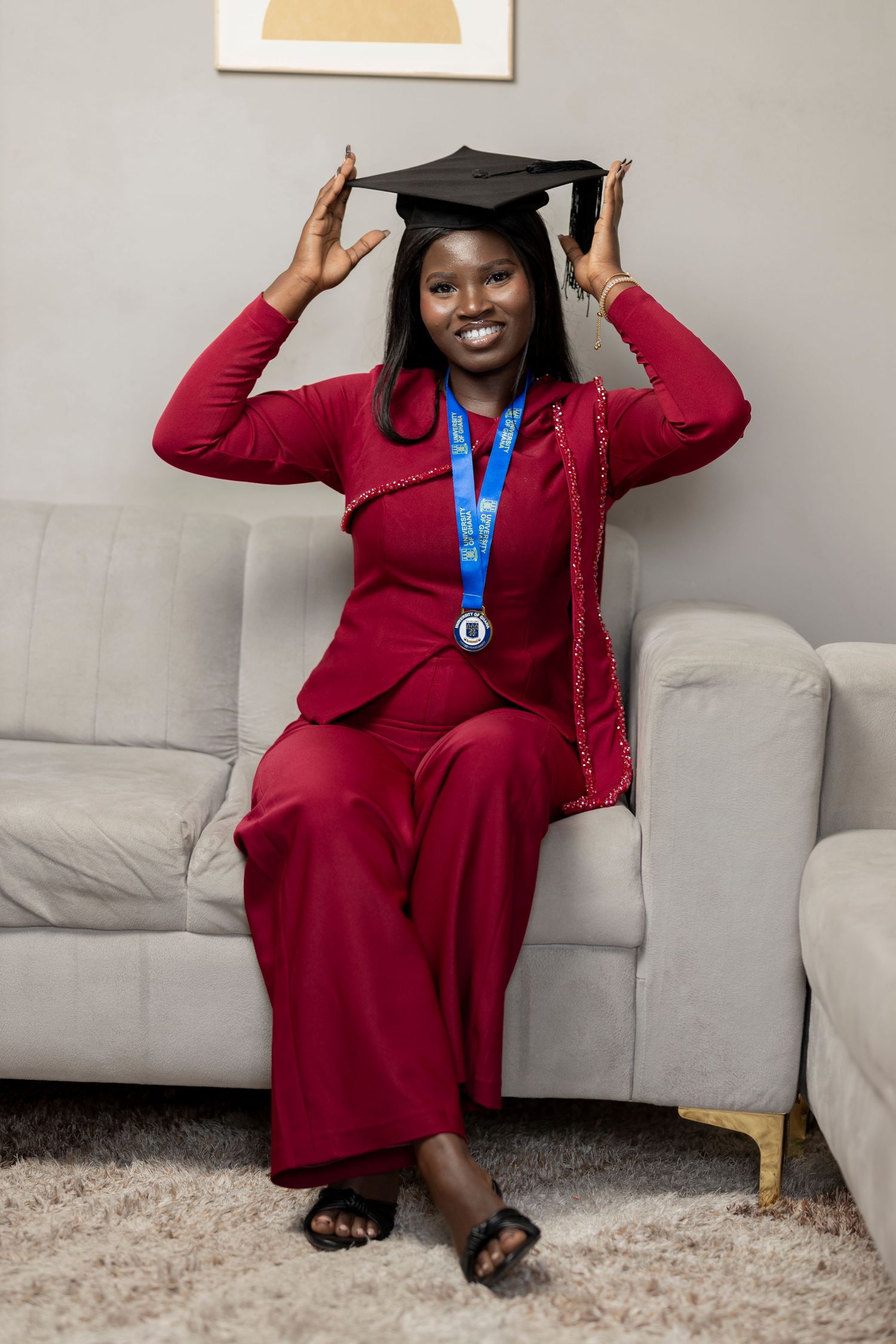 Smiling woman in red suit wearing graduation cap and blue medal, sitting on sofa celebrating academic achievement.