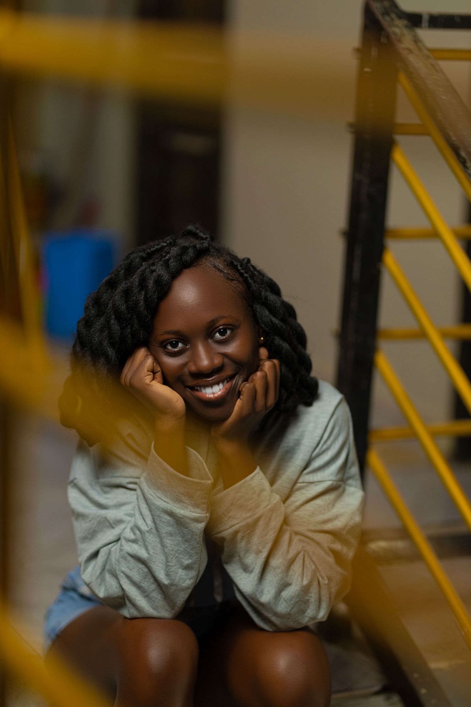 A joyful person in a gray sweater sits on stairs, framed through yellow railing in an indoor setting.