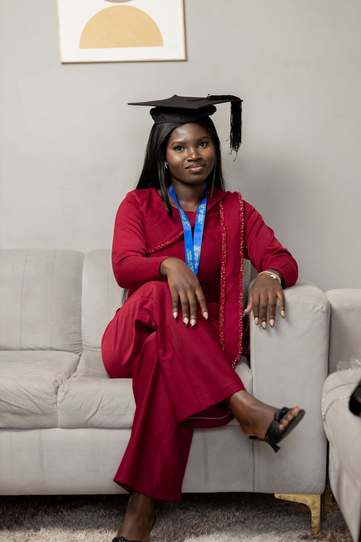 Young woman in red graduation gown and cap sits on white sofa, wearing blue honor cord, smiling proudly.