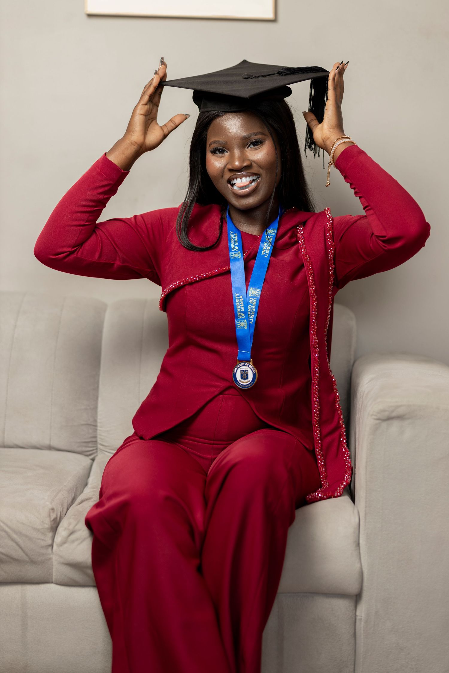 Smiling graduate in red suit wearing mortarboard cap and blue medal, sitting on sofa celebrating achievement.