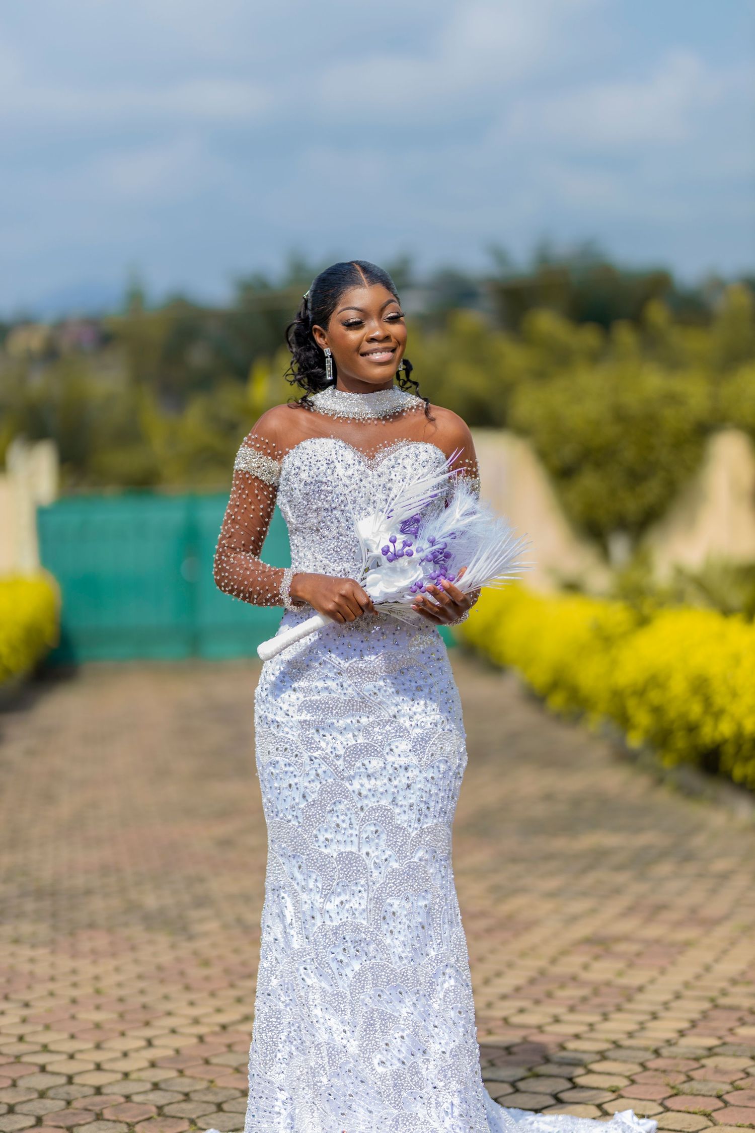 A stunning plus-size wedding dress with intricate lace details and off-shoulder design photographed in an outdoor garden setting.