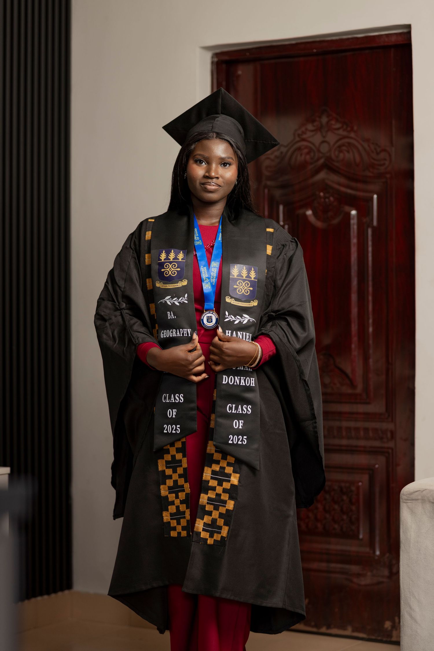 Young woman in black graduation gown, cap, and stole reading Class of 2025, posing proudly indoors.
