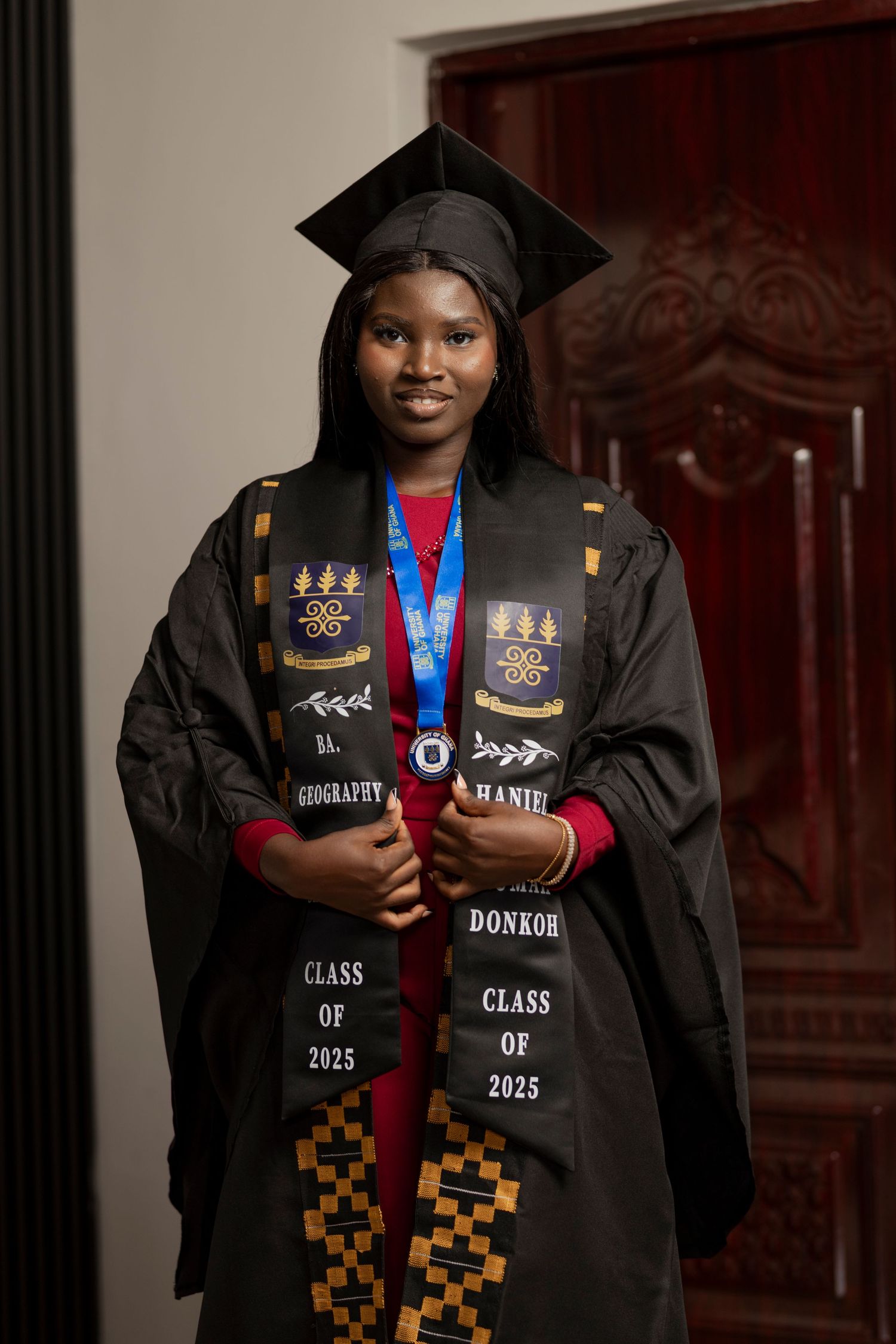 Young woman in black graduation cap and gown with Class of 2025 stole and medal, smiling indoors.