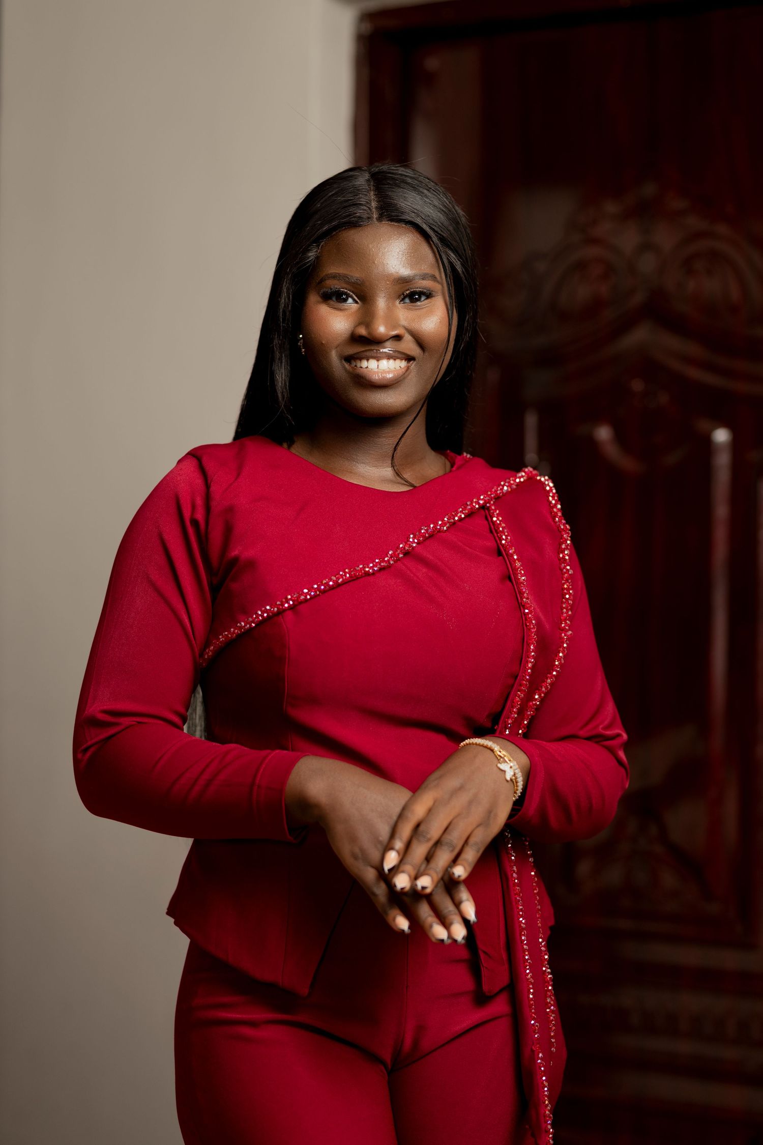 Young woman smiling in a red long-sleeve outfit with gold chain detail, posing indoors.