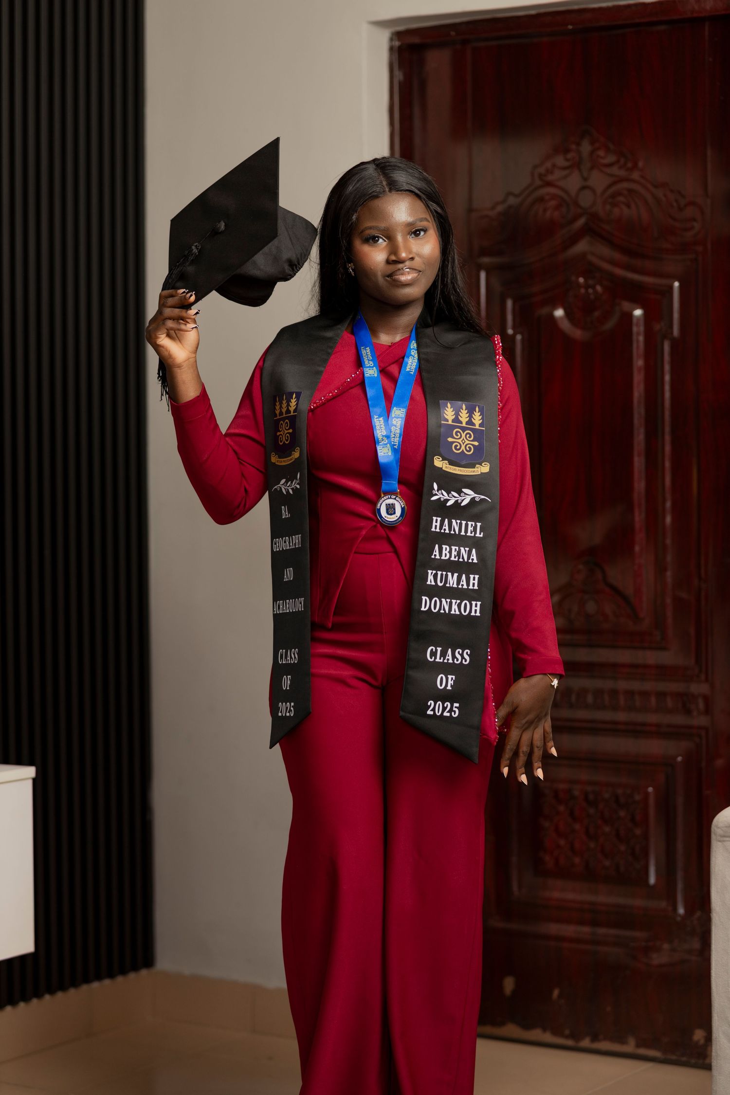 Graduate Haniel Arena Kumar Donkoh poses in red outfit with Class of 2025 stole, medal, and mortarboard cap.