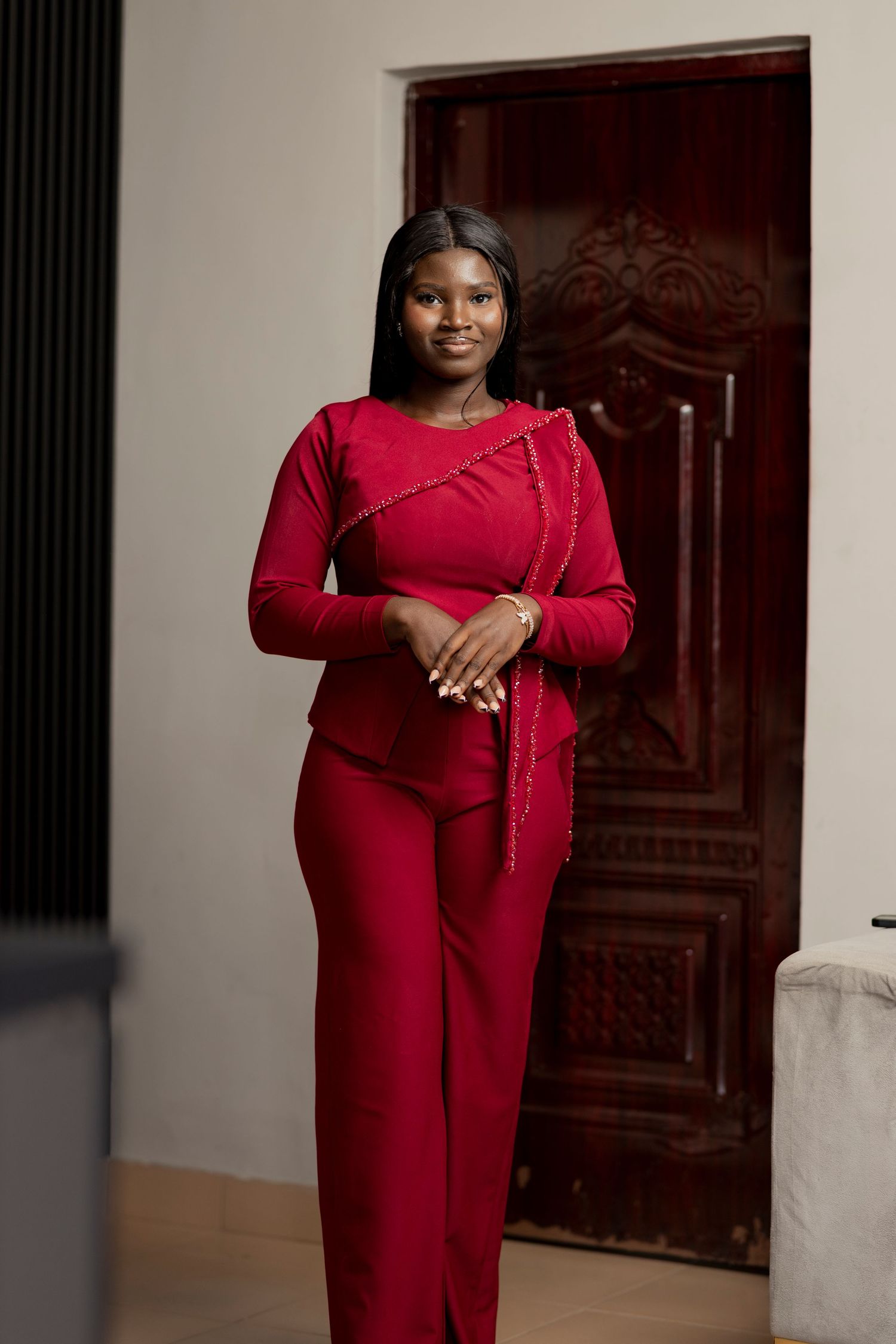 Woman in elegant red outfit with gold trim stands confidently near ornate wooden door.