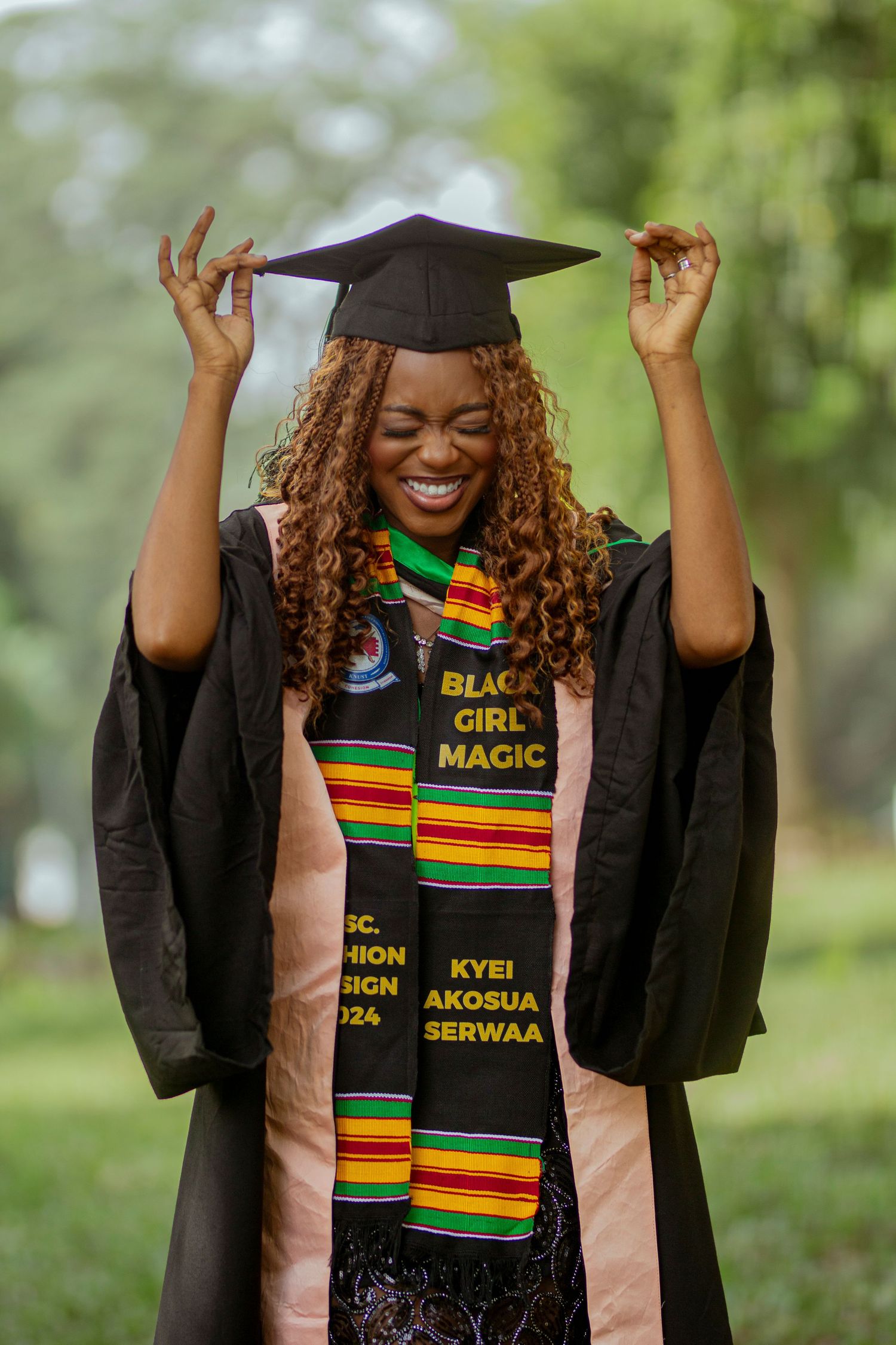 Graduate wearing black cap and gown with colorful kente stole celebrates achievement in outdoor setting.