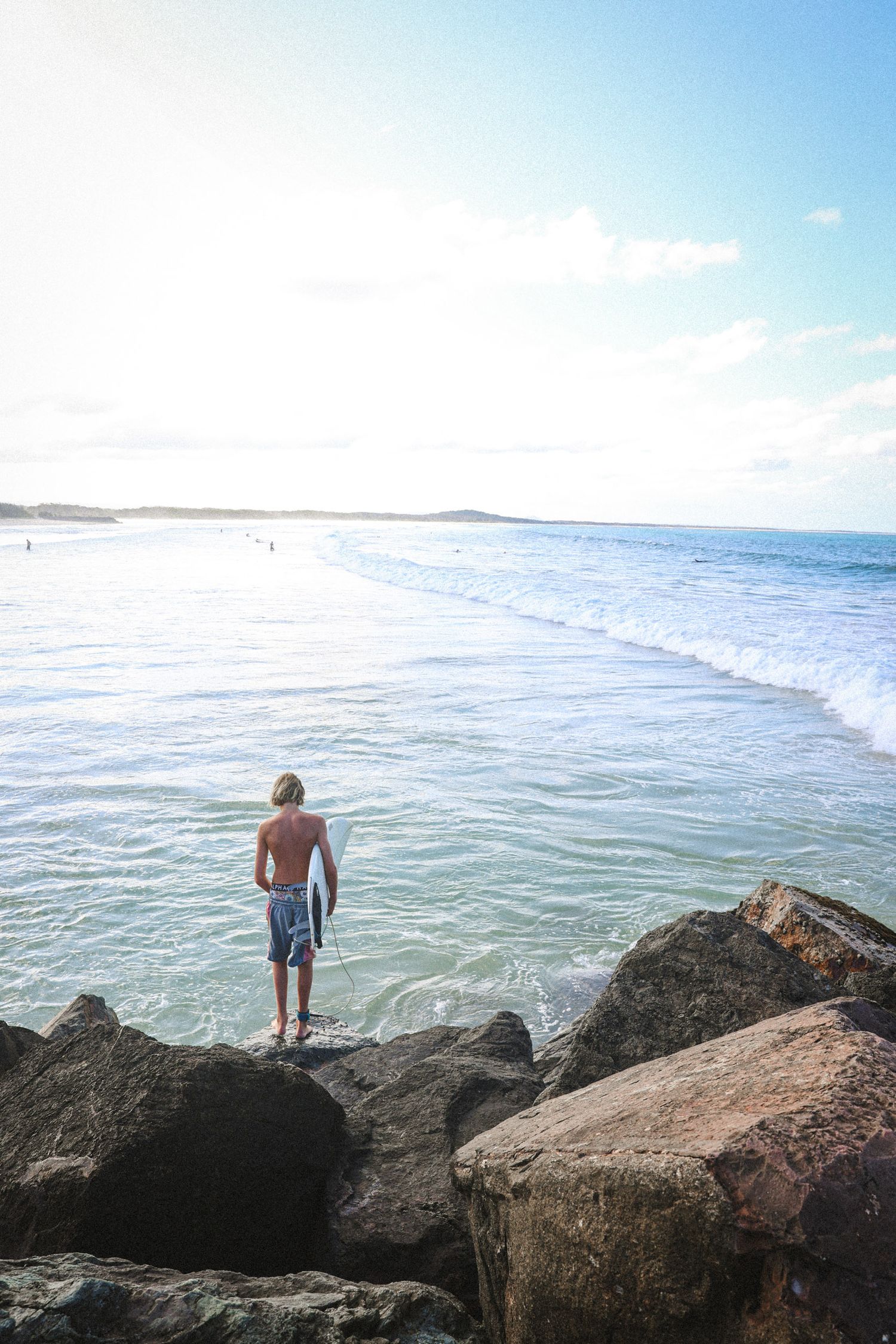 Figure standing on rocky outcrop overlooking ocean waves at sunny beach.