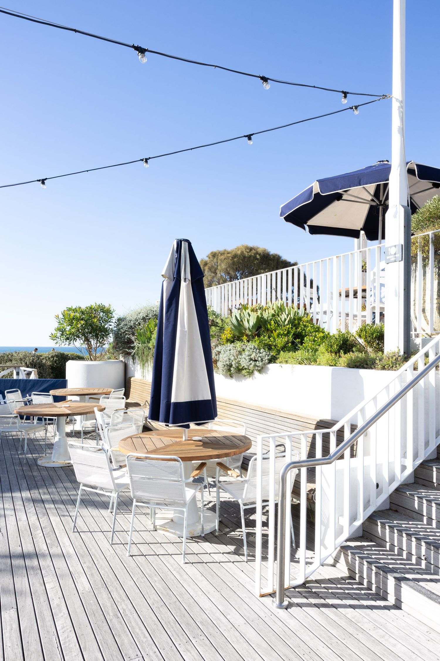 Elegant outdoor dining deck with string lights, navy umbrellas and white railings overlooking the ocean.