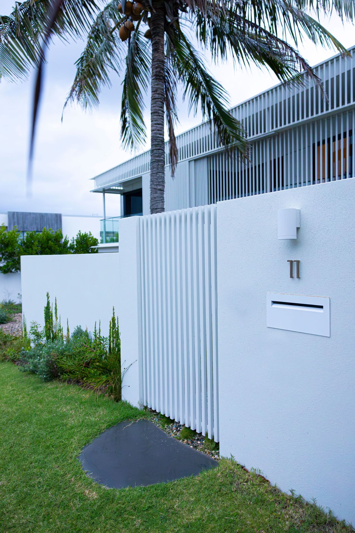 Modern white house exterior with vertical slats and palm trees against cloudy sky.