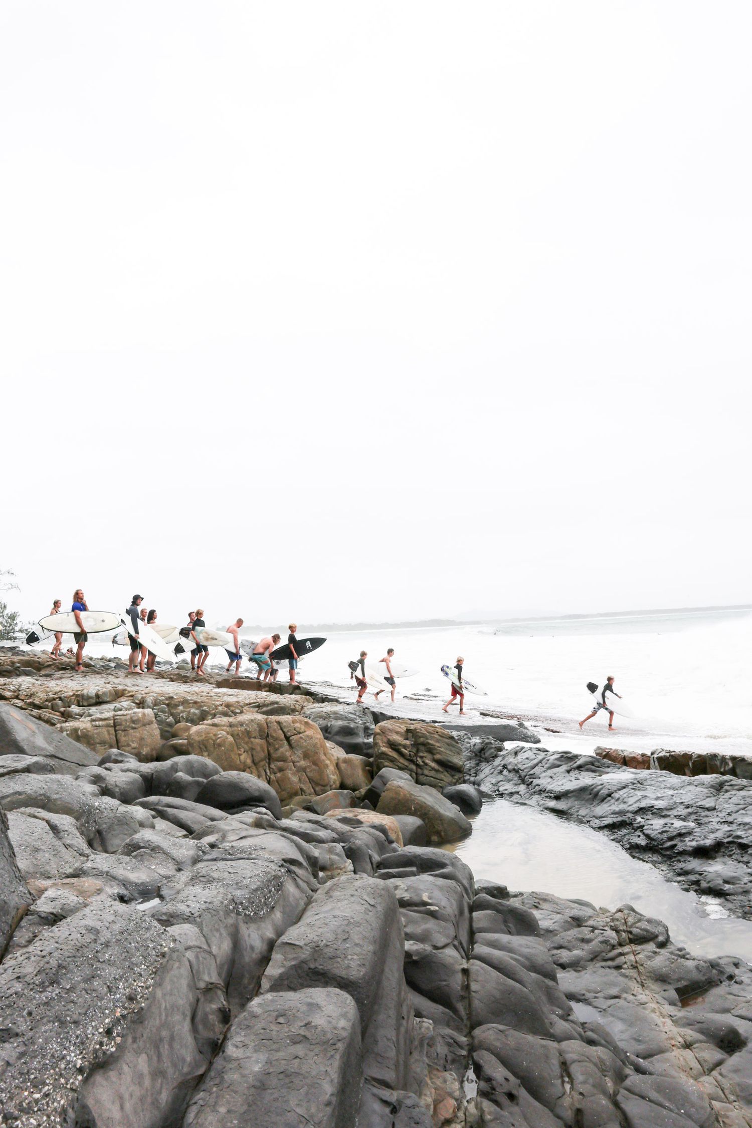 Crowd traverses rocky coastal formation with surfboards in overcast weather.