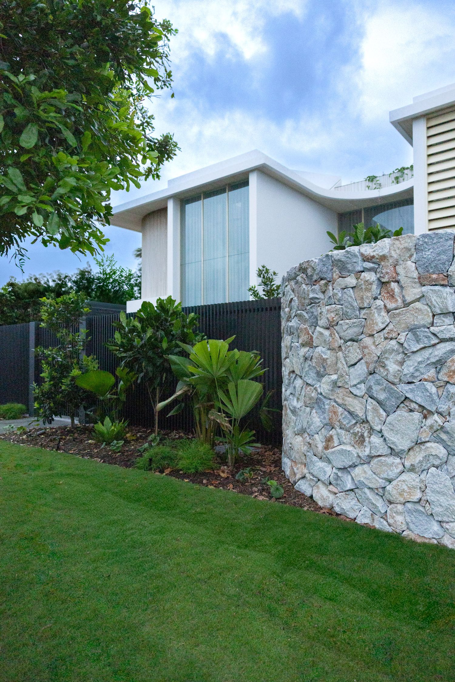 Modern white house exterior with stone wall feature and lush tropical landscaping.