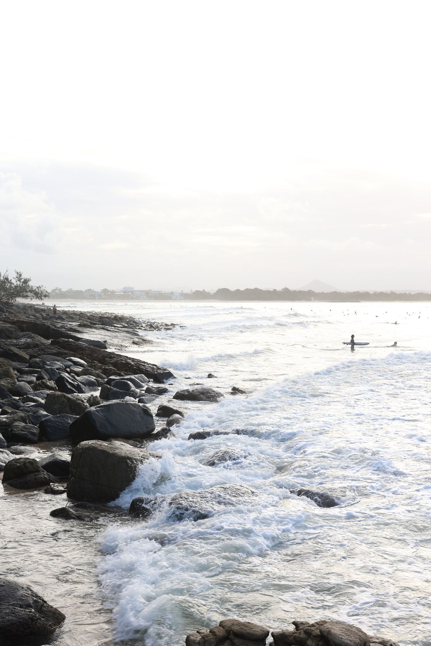 Rocky coastline with crashing waves and distant surfers at dusk.