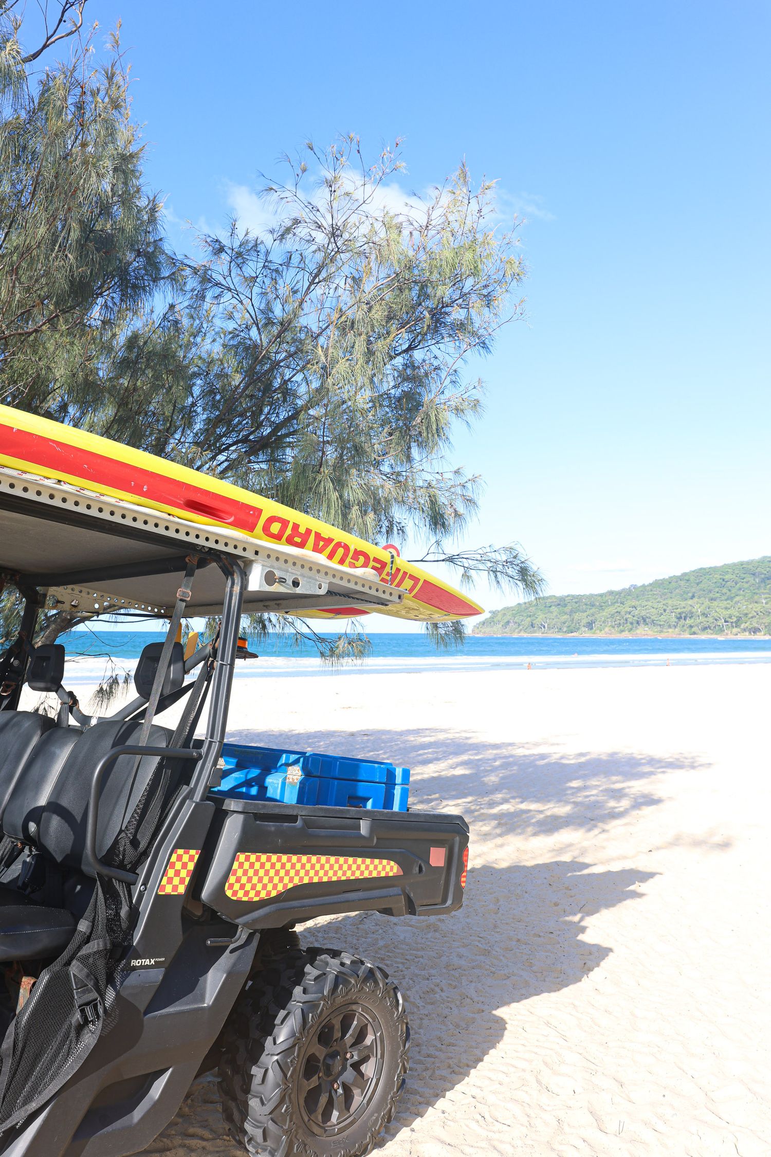 Off-road vehicle with surfboard on top parked on pristine beach with blue waters in background.
