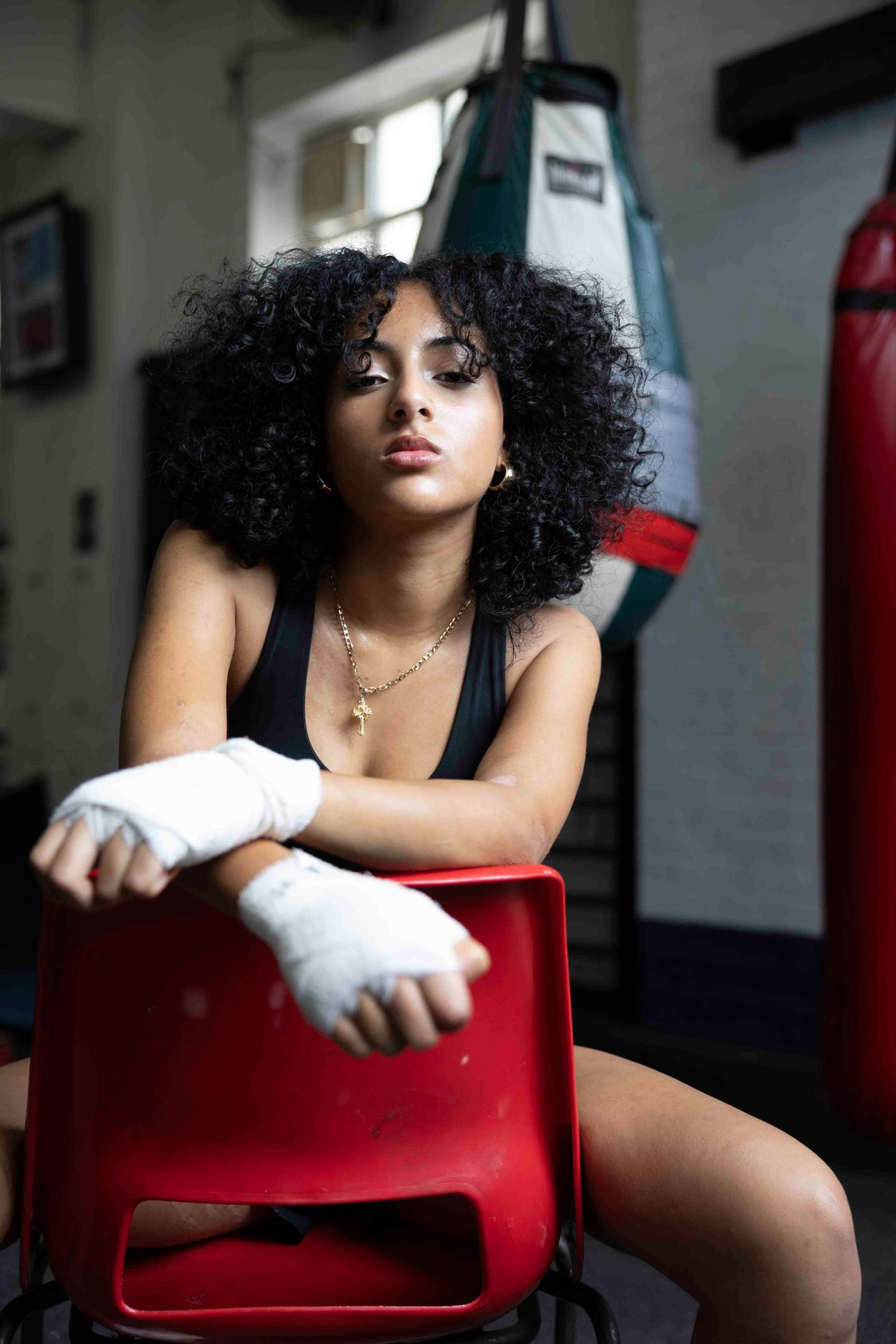 Athletic figure with curly hair poses confidently in boxing gym wearing black tank top and hand wraps.