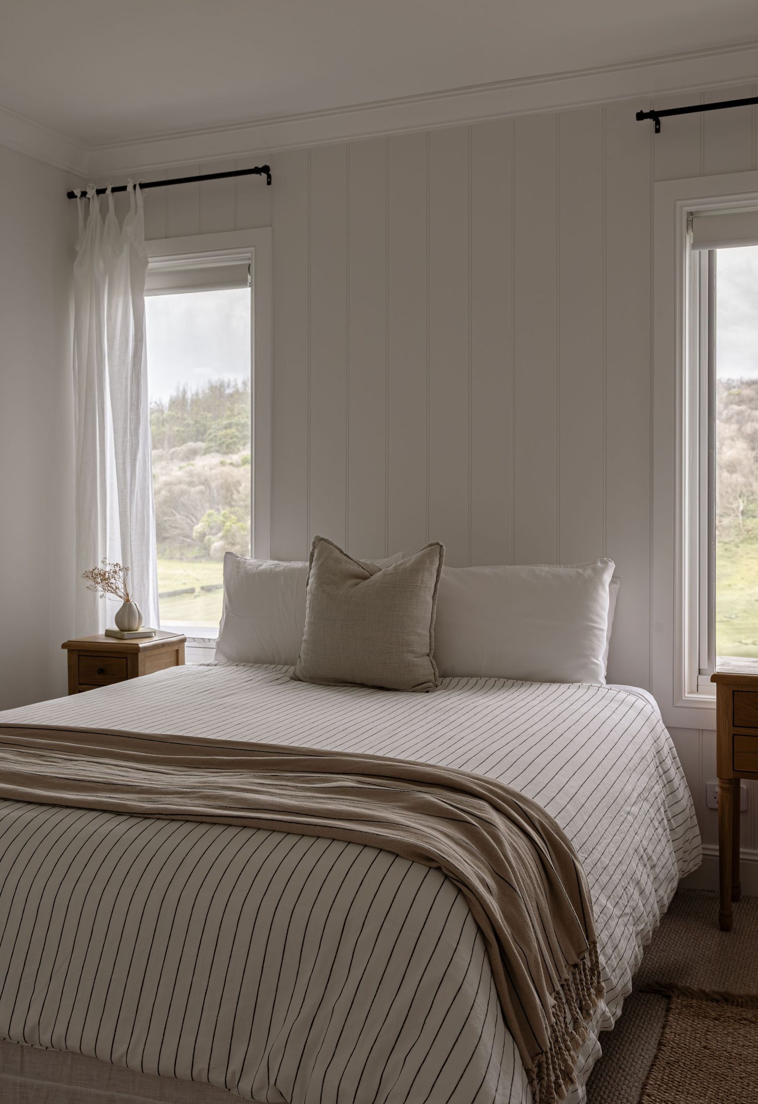 Serene bedroom with white linens, natural light from two windows, and a cozy brown throw blanket.