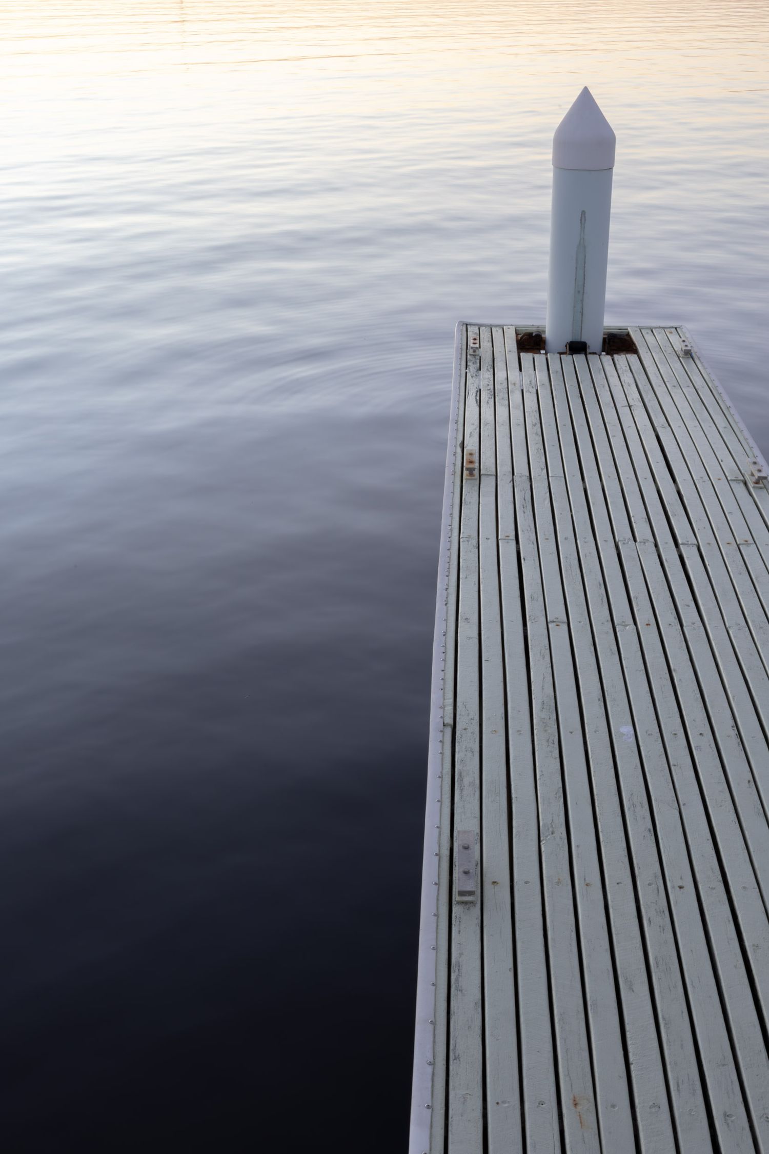 Minimalist wooden pier with white post extends into still waters during twilight.