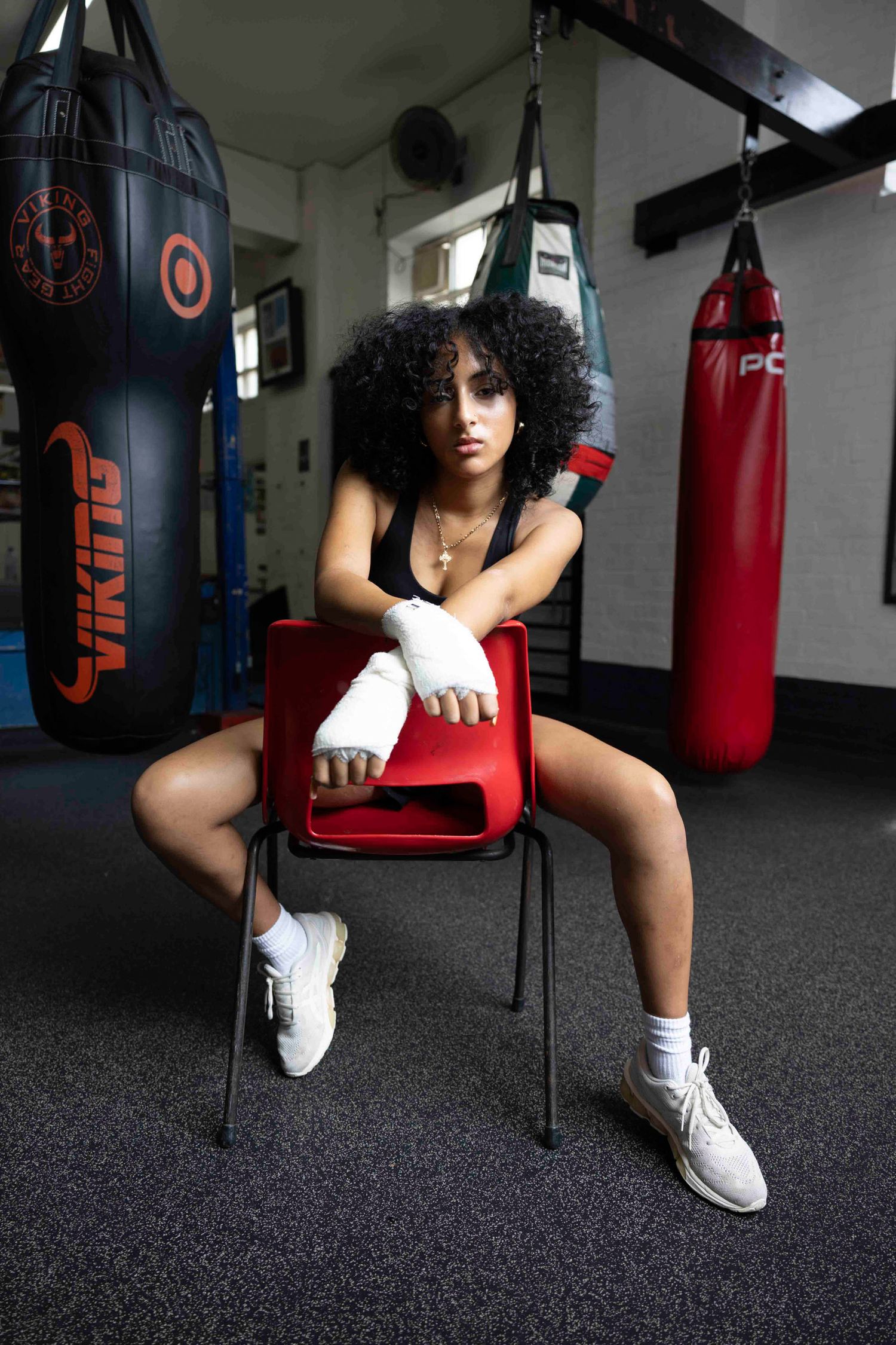 Boxer sits on red chair in gym surrounded by punching bags wearing athletic attire.