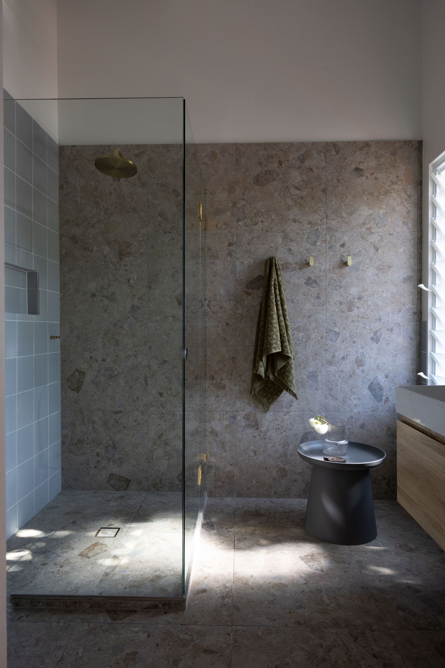 Modern minimalist bathroom with glass shower partition, stone walls, and hanging towel in natural sunlight.