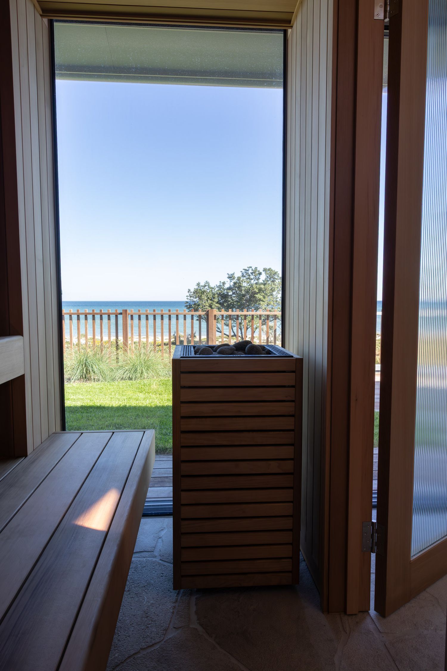 View from inside a sauna looking out through glass doors to beach and ocean with wooden bench and slat box in foreground.