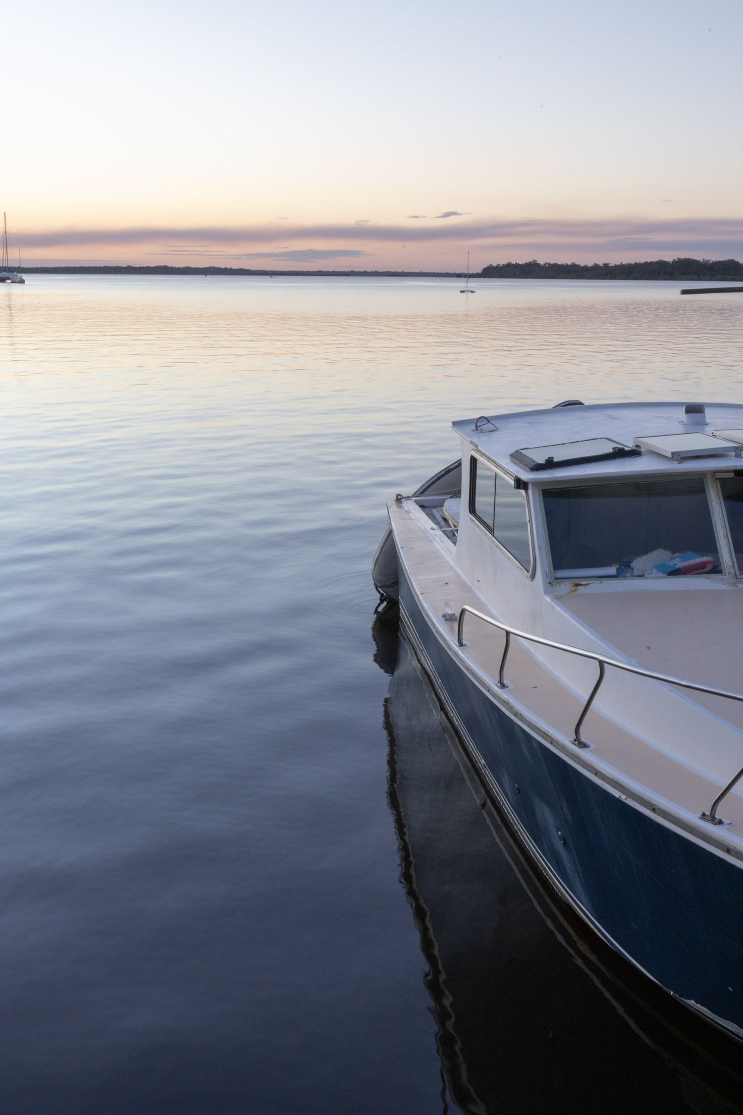 Boat moored at calm harbor during sunset with pastel sky reflections on still water.