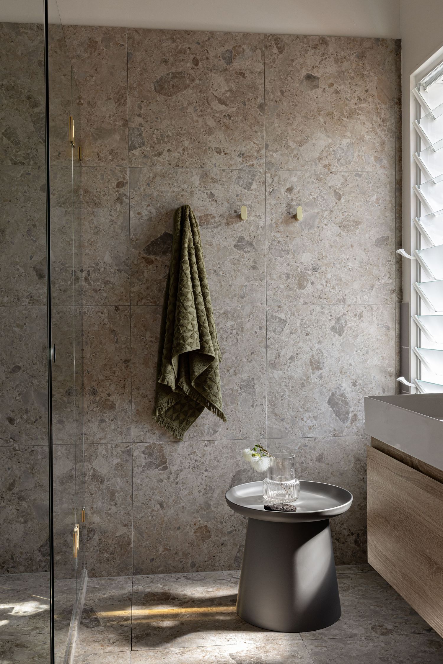 Modern bathroom interior with textured concrete walls, glass shower partition, and black side table holding decorative items.