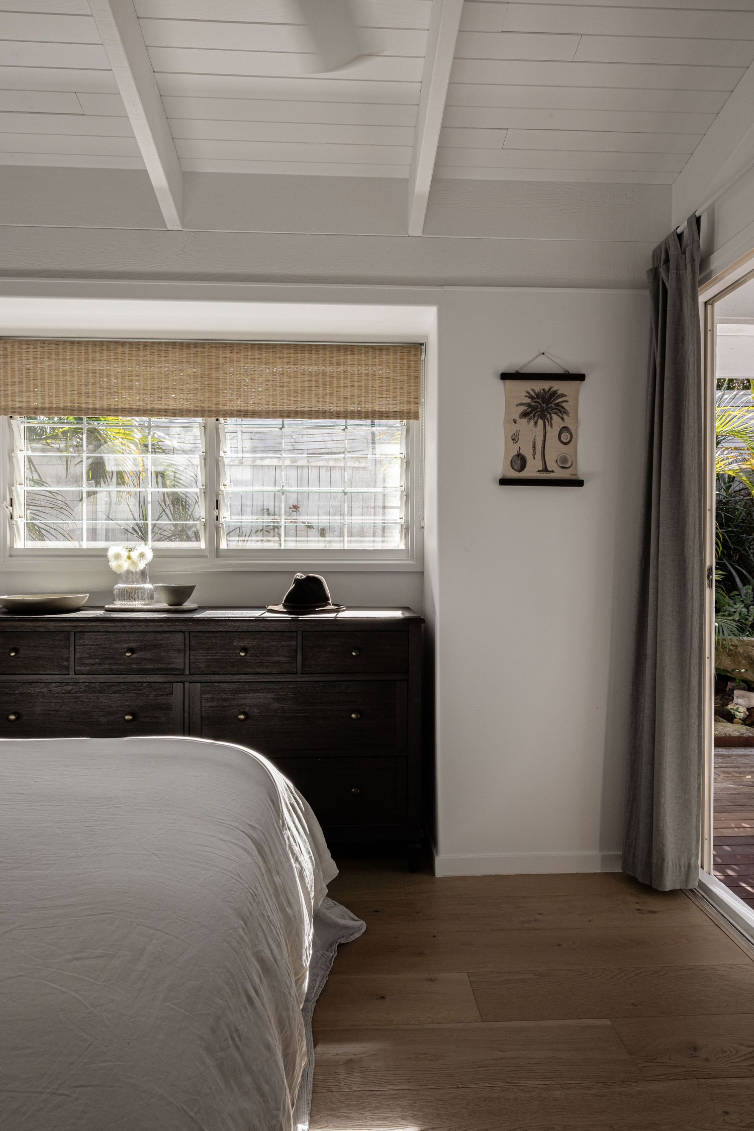 Modern bedroom interior with white walls, dark wood flooring, and natural light streaming through horizontal windows.