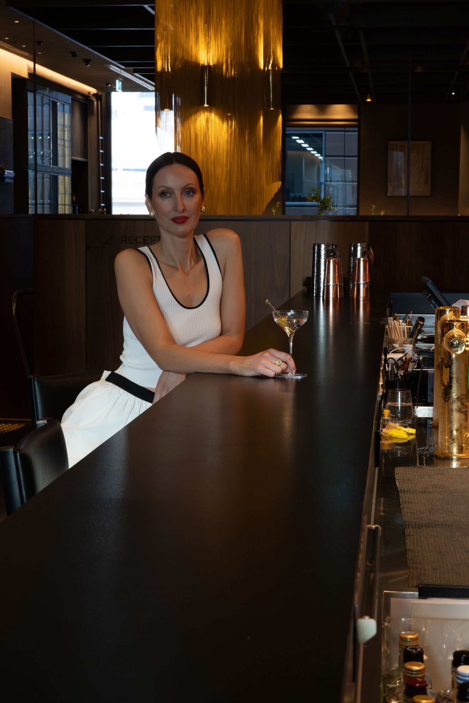 Elegant woman in white dress sitting at a sleek dark bar with a martini glass in an upscale restaurant.