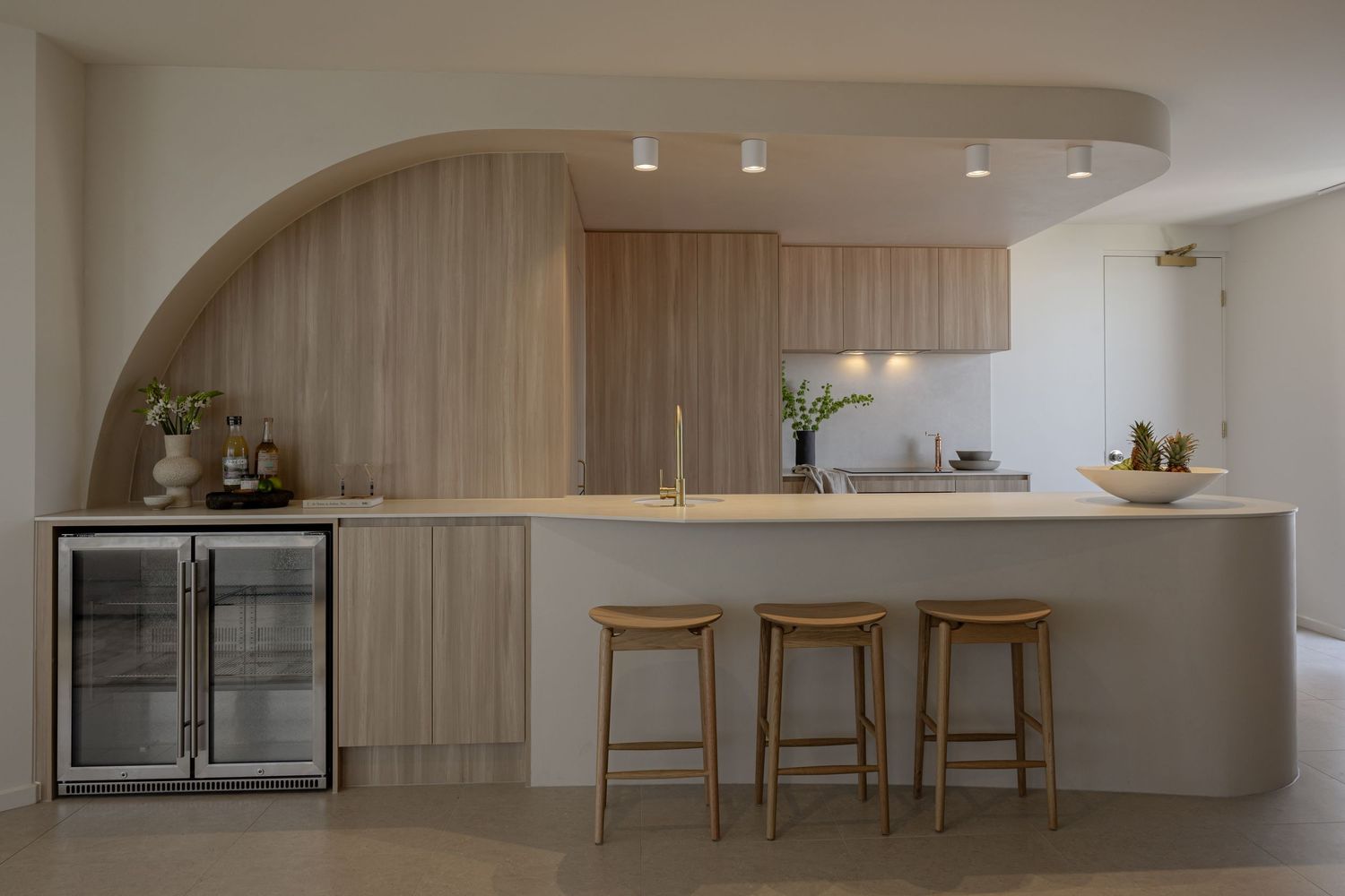 Modern kitchen with curved wood paneling, white countertops, bar stools, and glass-door beverage fridge in neutral tones.