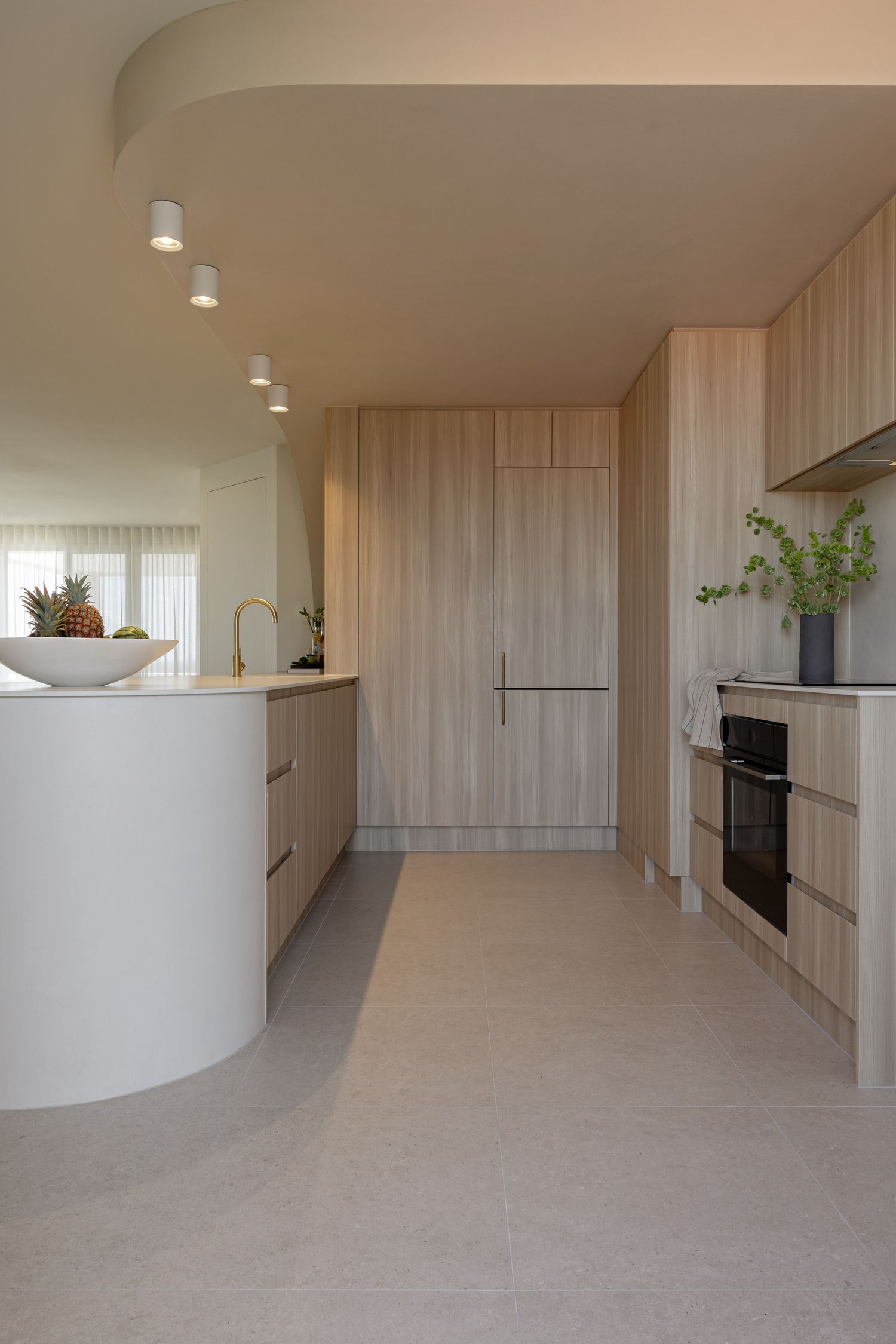 Modern minimalist kitchen with curved white walls, light wood cabinets, and track lighting in an open-plan layout.