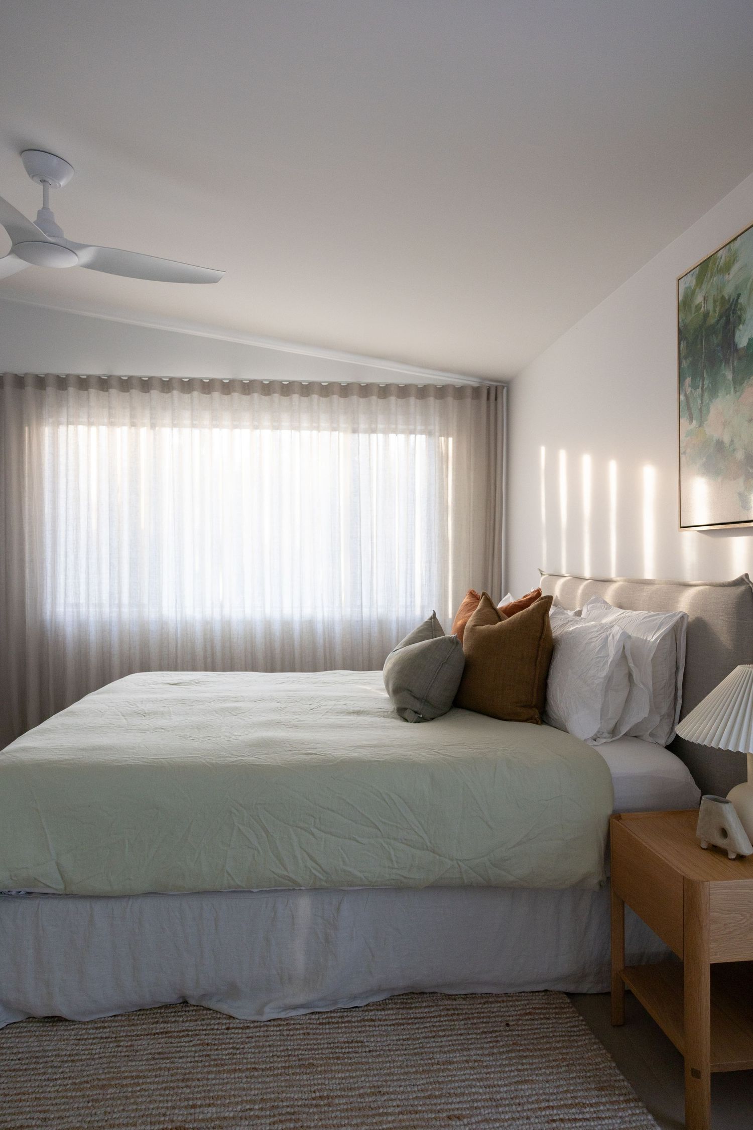 Simple bedroom interior with white bedding, wooden furniture, and sheer curtains filtering natural light through windows.