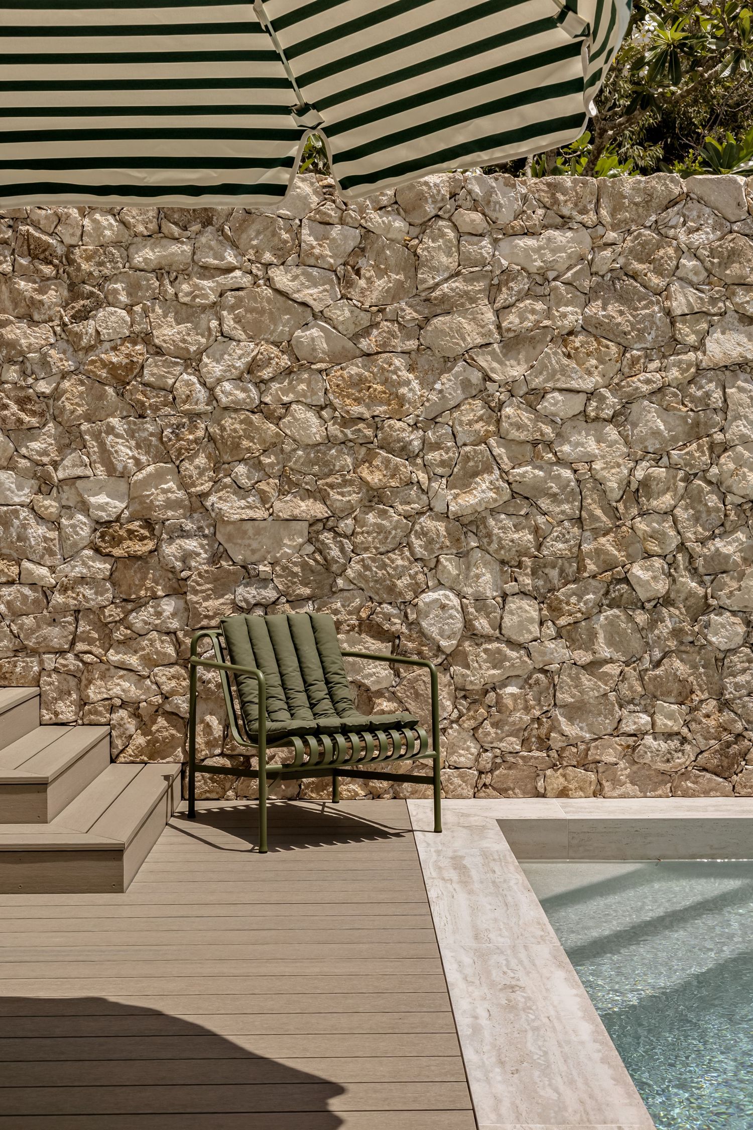 Green lounge chair beside a stone wall and small pool on a wooden deck under a striped umbrella.