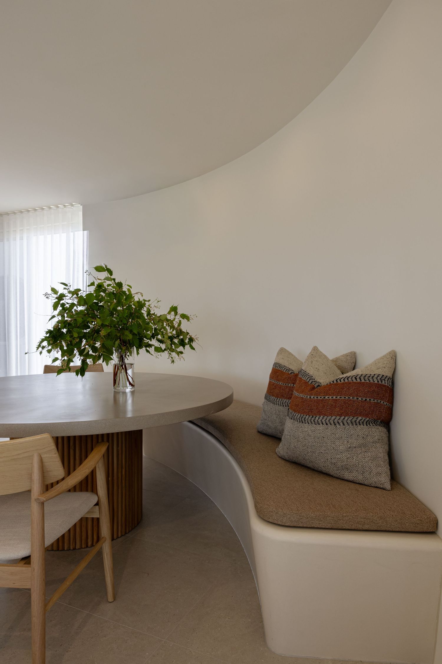 A minimalist curved seating area with white built-in benches, wooden accents, and decorative pillows in a bright room.