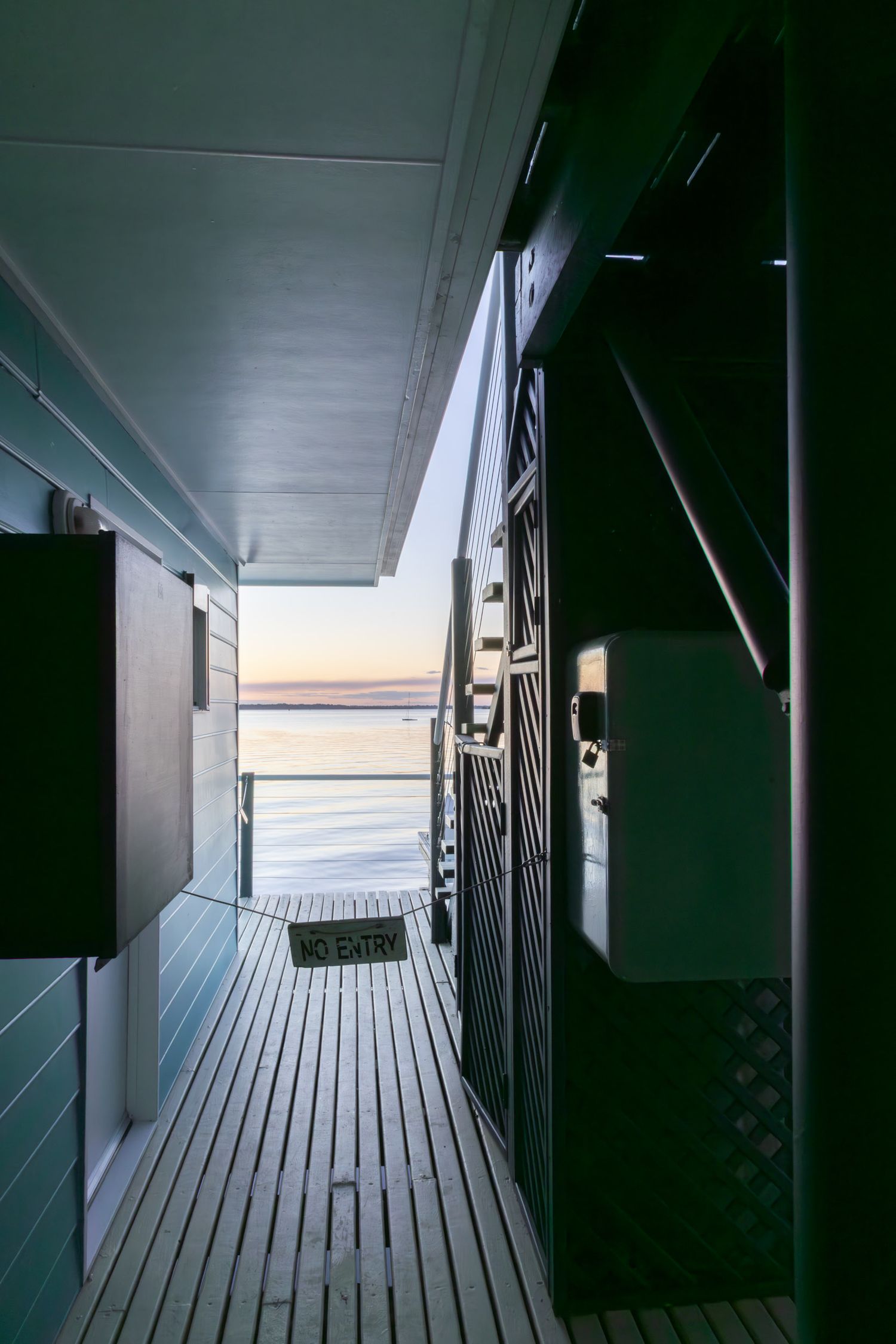 Narrow wooden boardwalk corridor between boat storage units with water view at dusk.