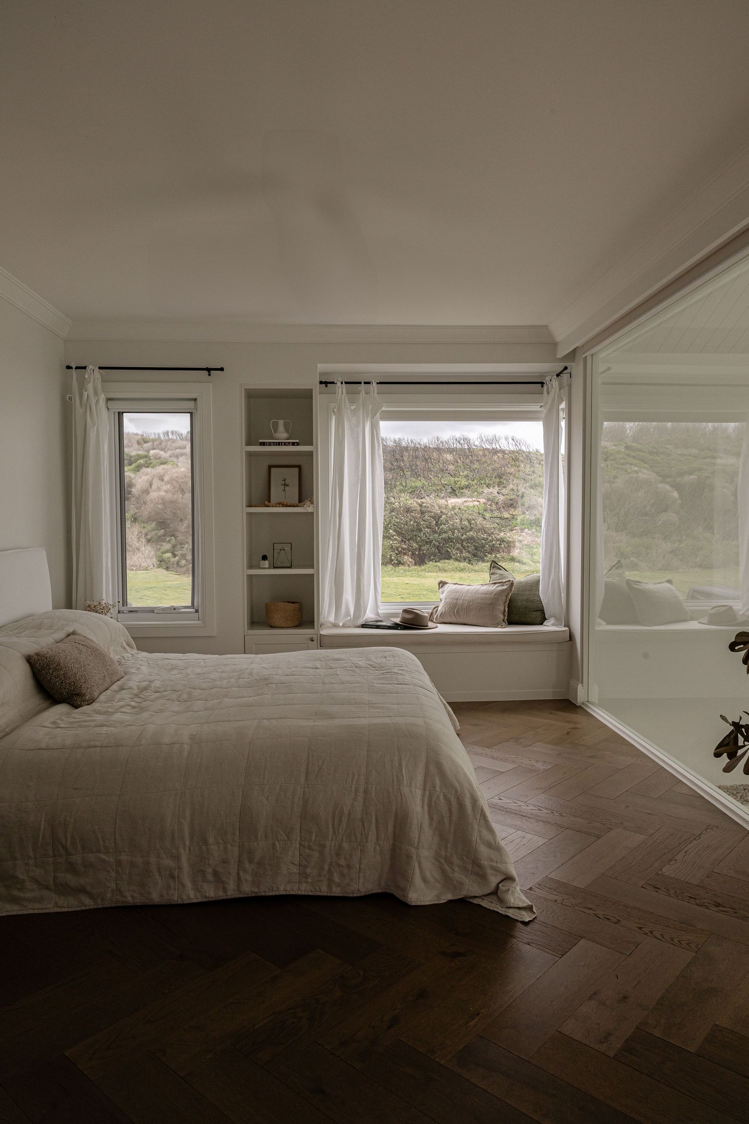 Minimalist bedroom interior with panoramic windows, dark hardwood floors, and neutral bedding in white tones.