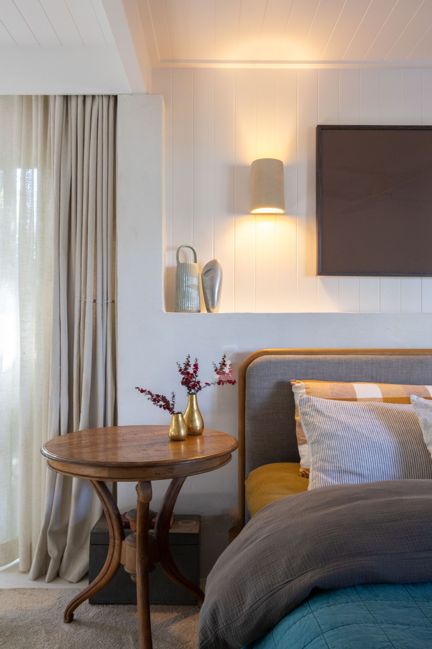 Cozy bedroom interior with round wooden side table, wall sconce, and gray bedding against white paneled walls.