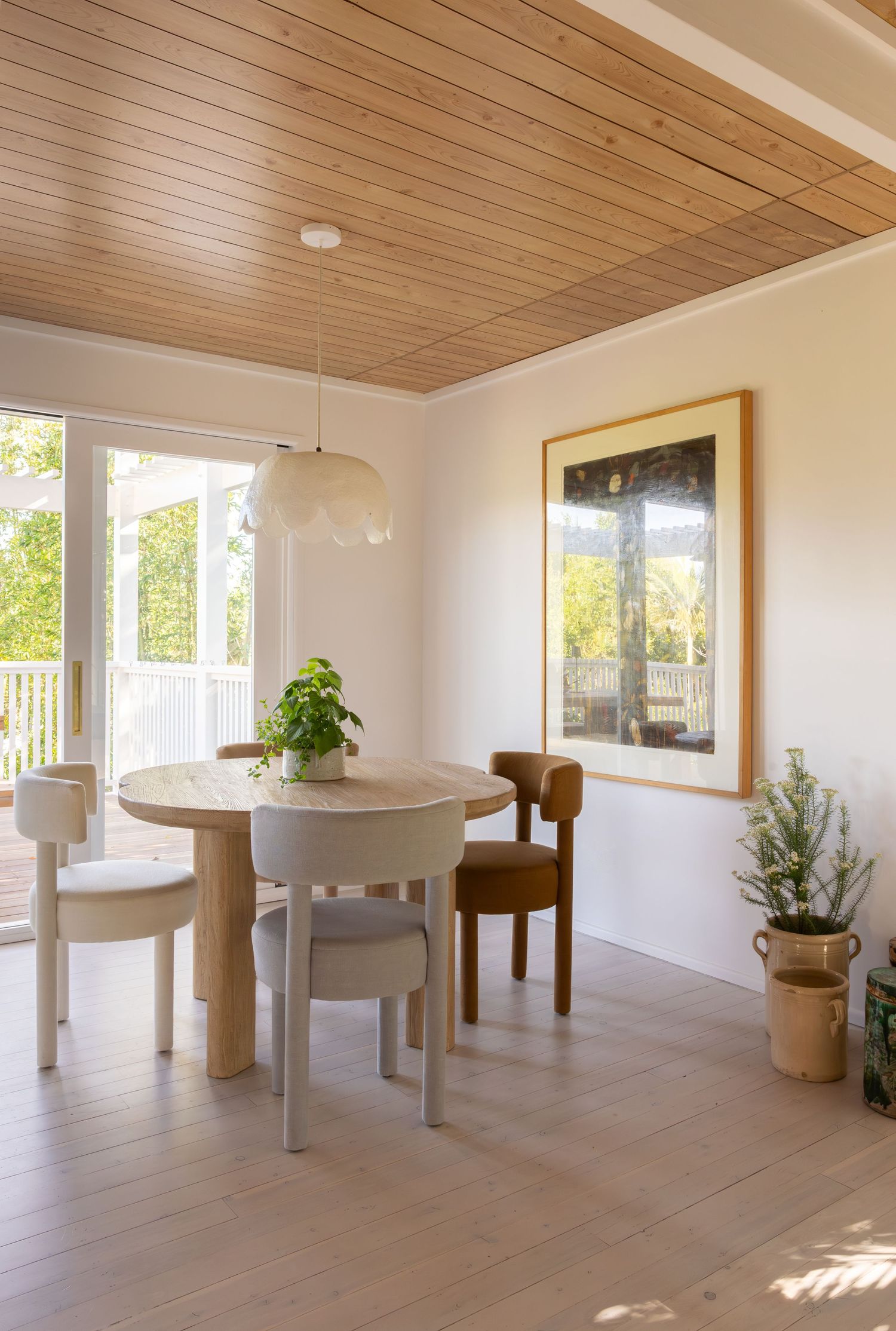 Modern sunlit room with wood ceiling, light hardwood floors, round white tables, and large windows overlooking outdoor space.