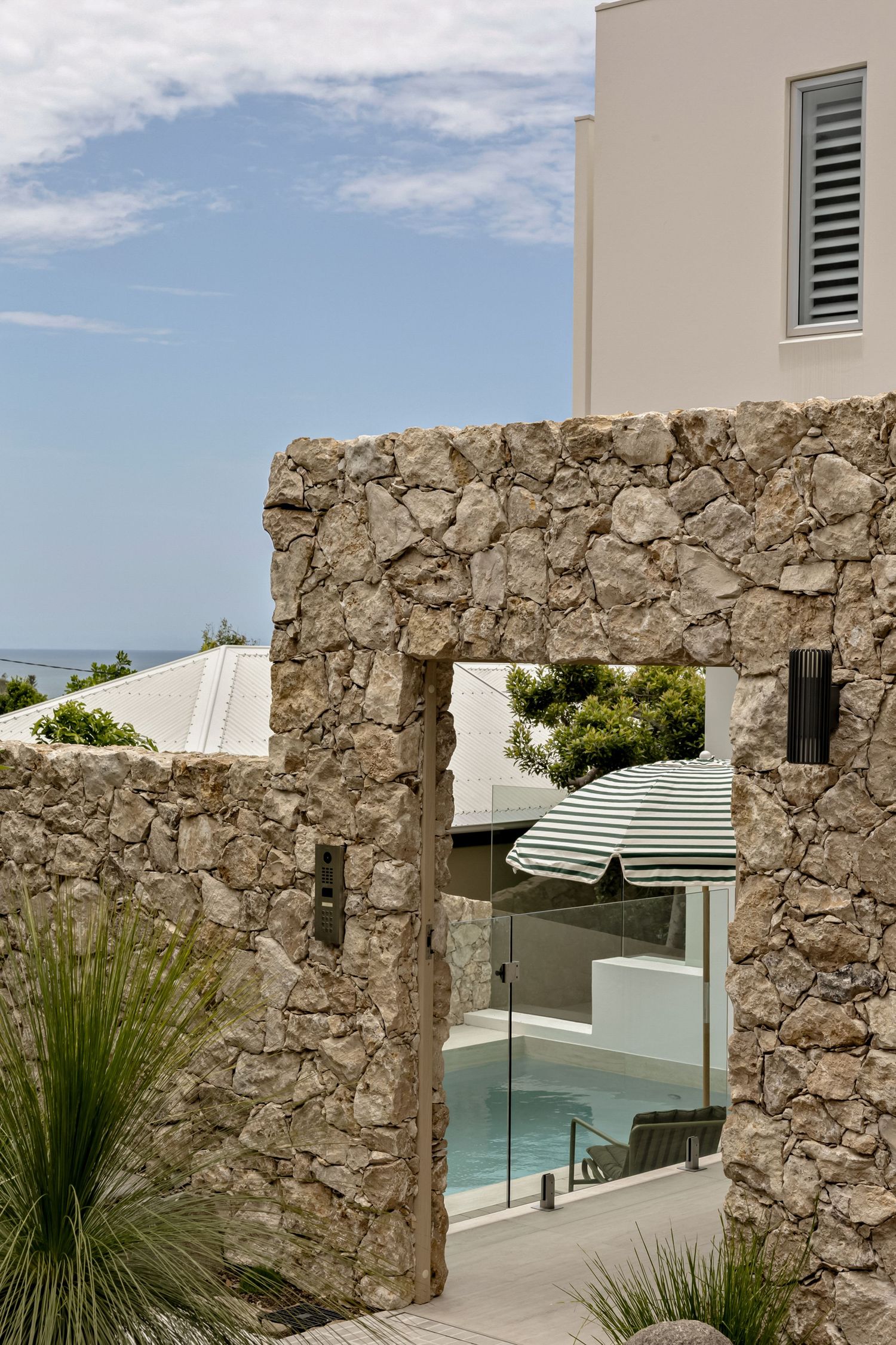 Stone archway entrance leading to a modern coastal home with a pool and striped umbrella, ocean views in the background.