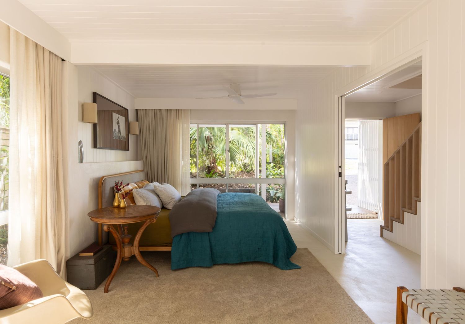 Modern bedroom with teal bedding, wooden furniture and large windows overlooking tropical garden views.